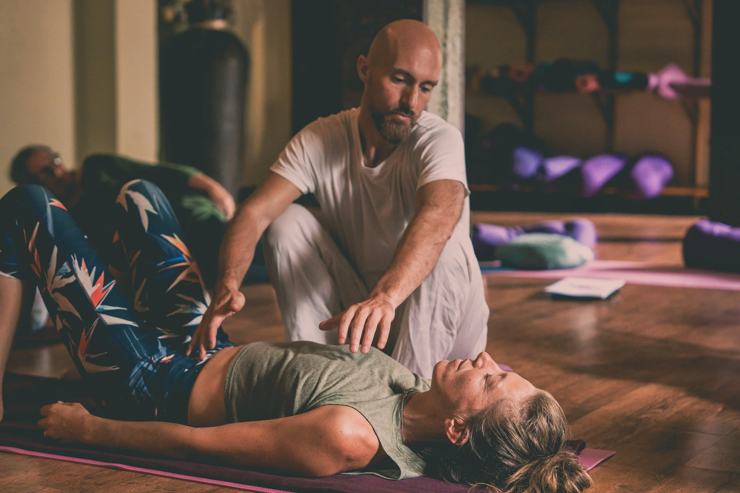 A man practicing CPR on a woman lying on a yoga mat, with another person in the background lying on a mat, in a room equipped for fitness or yoga.