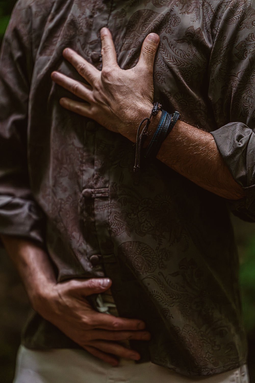 Person with one hand on his chest and the other on his stomach, wearing a patterned shirt and bracelets on his wrist.