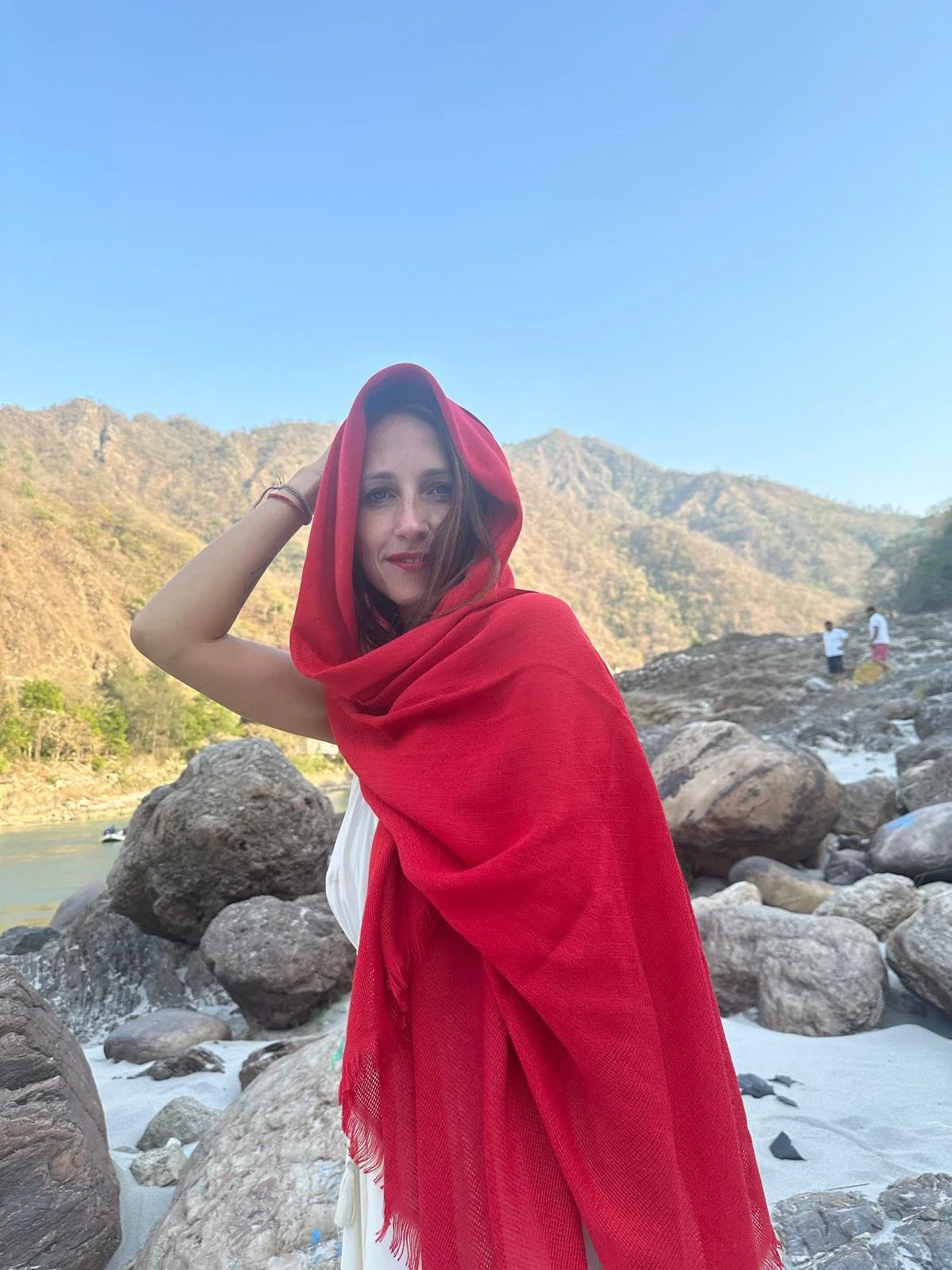 A woman with a red shawl over her head standing on rocks near a river with mountains in the background.