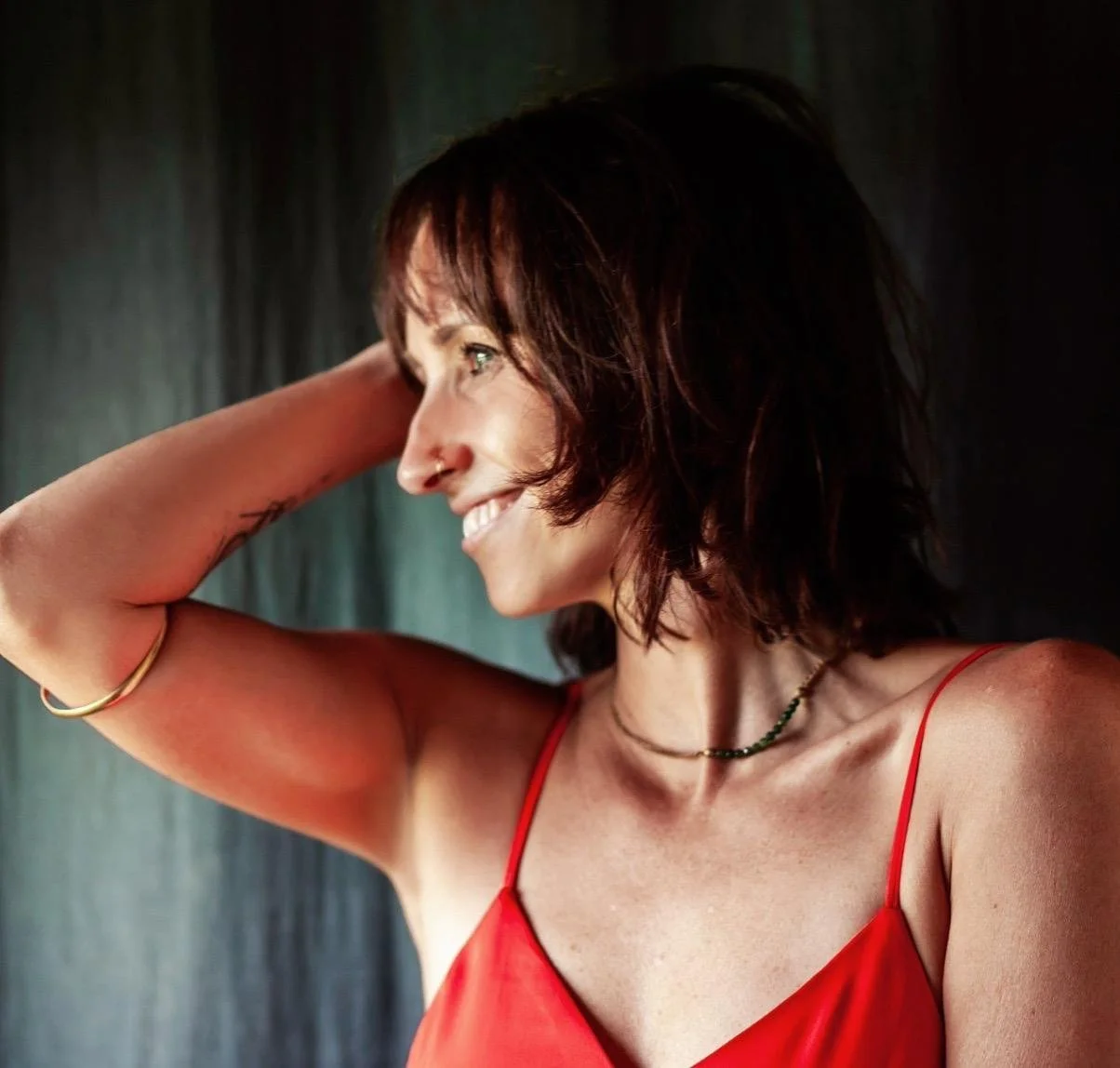 A woman in a red spaghetti strap top, smiling and looking to the side, with short brown hair and jewelry, standing against a dark background.