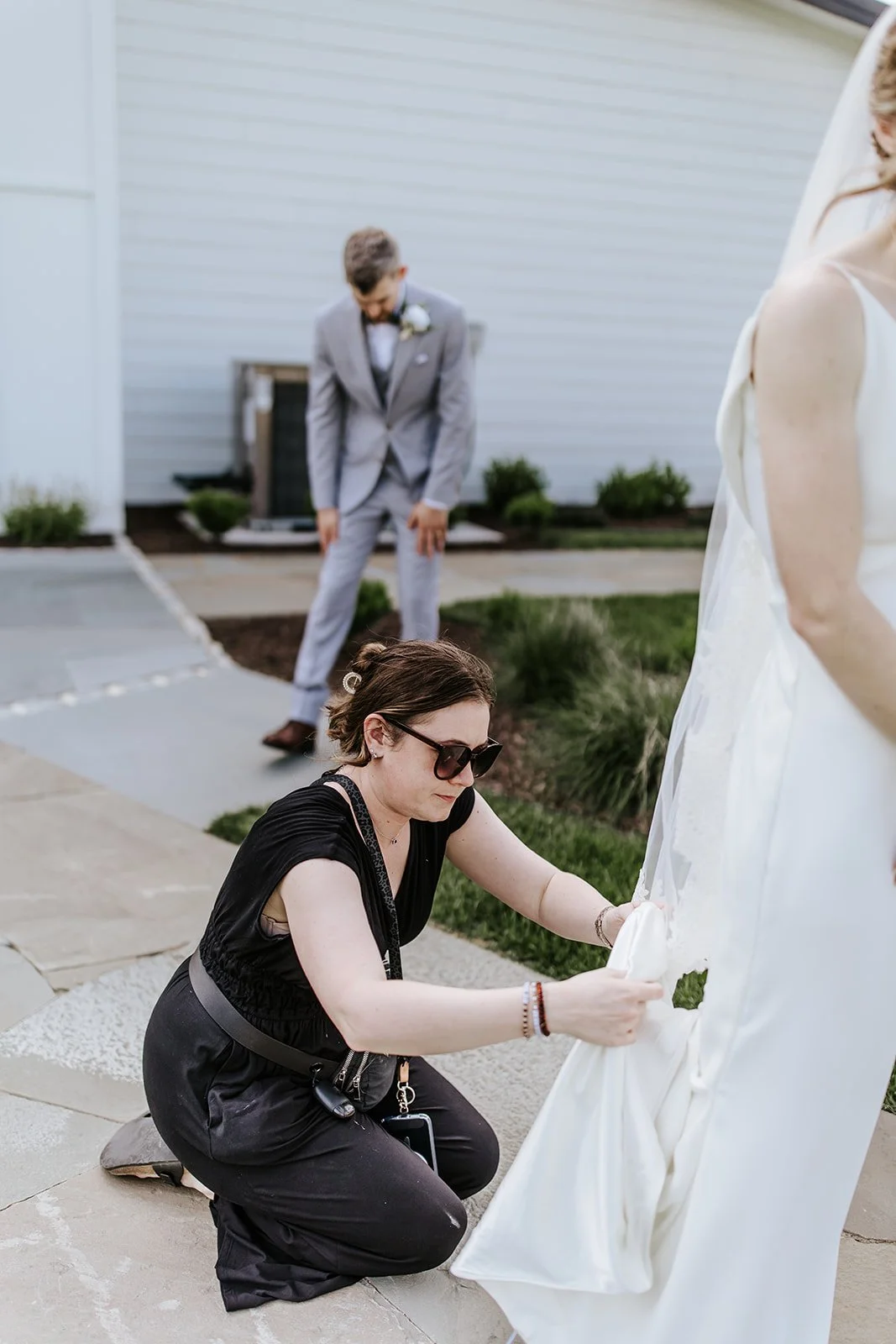 A woman adjusting her wedding dress outside, with a man in a gray suit in the background.