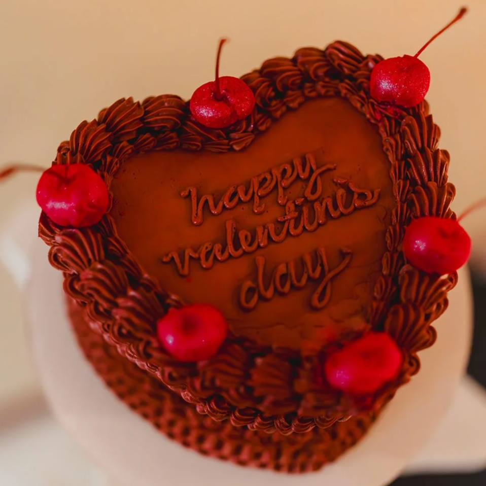 Chocolate heart-shaped cake with chocolate frosting, topped with five red cherries, with a message that reads "happy valentine's day."