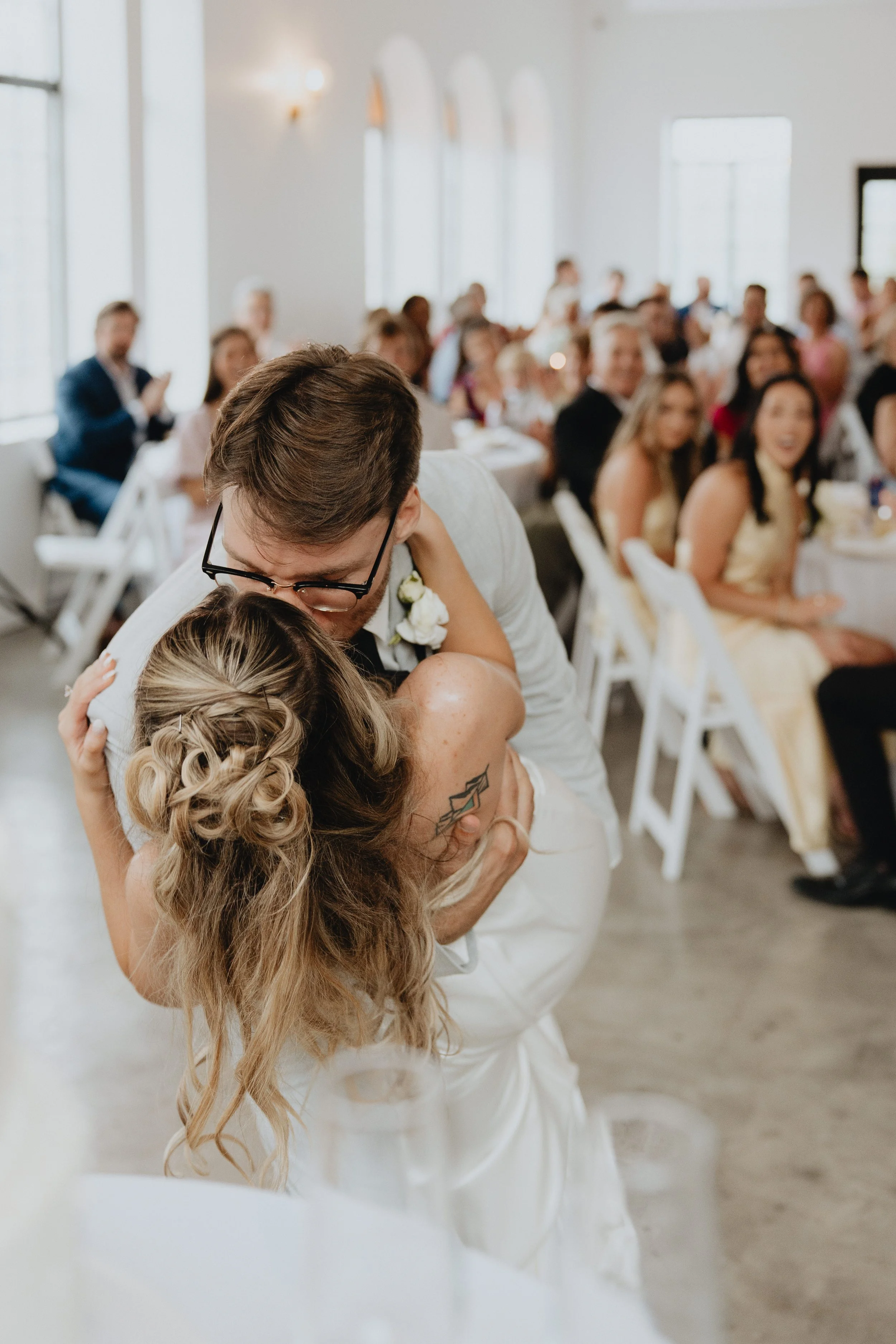 Couple hugging and kissing during a wedding reception with guests seated at tables in the background.