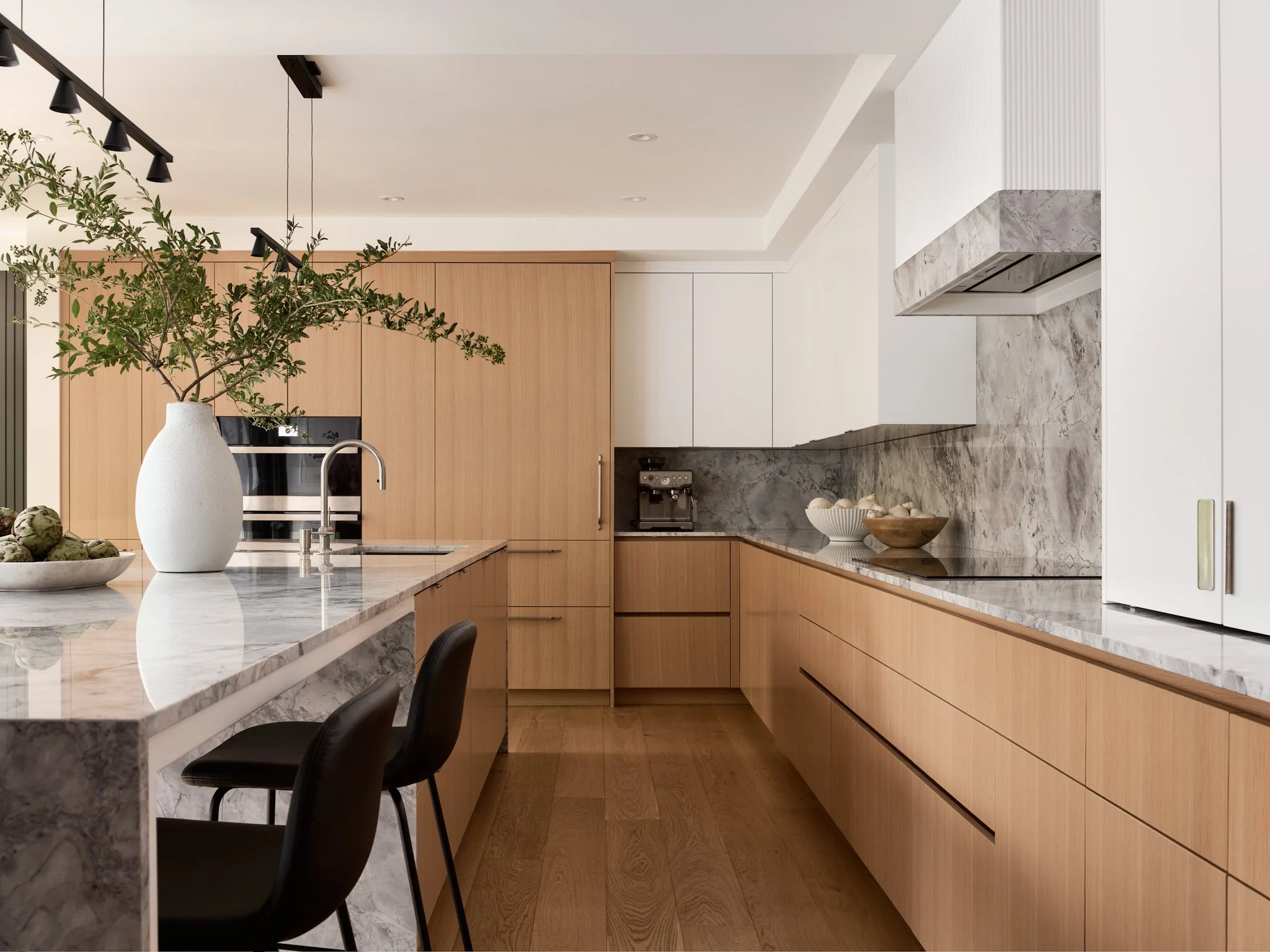 A modern kitchen with white cabinets, a dark gray island, brass hardware, a gold faucet, and a black range hood. There are decorative items like a cutting board, a bunch of herbs, and a utensil holder on the countertops. A small rug is on the wooden floor.
