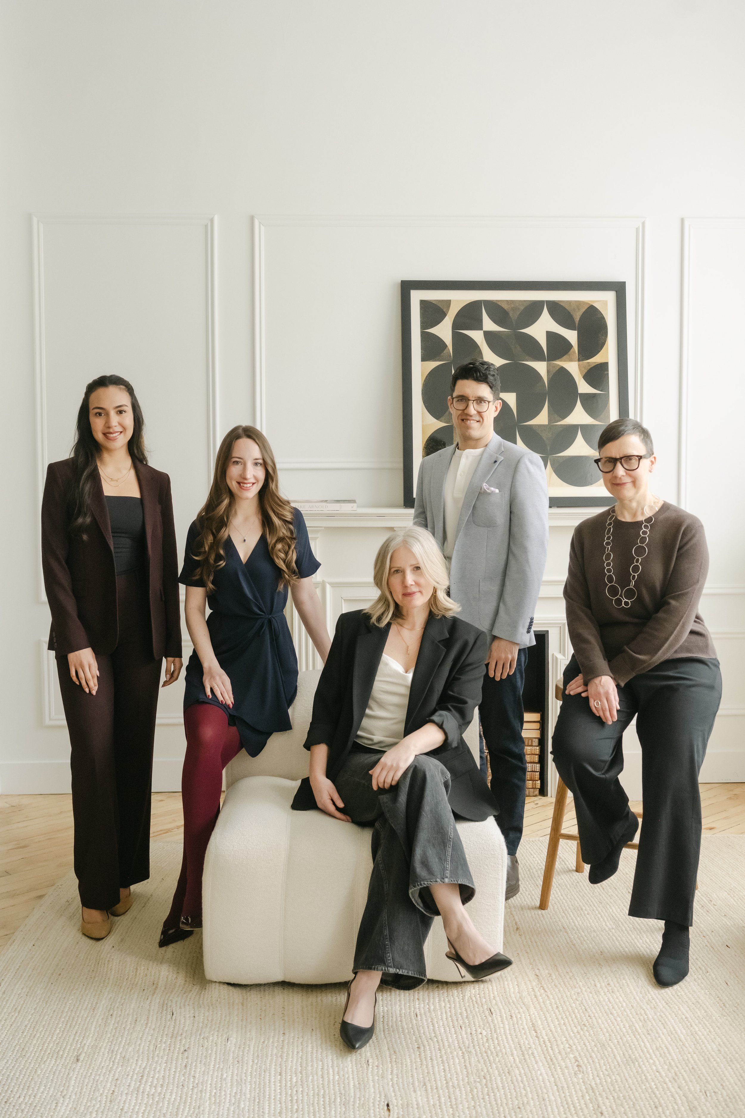 Group of five diverse people in business casual attire in a bright, airy room with white curtains and hardwood floors, posing for a photo.