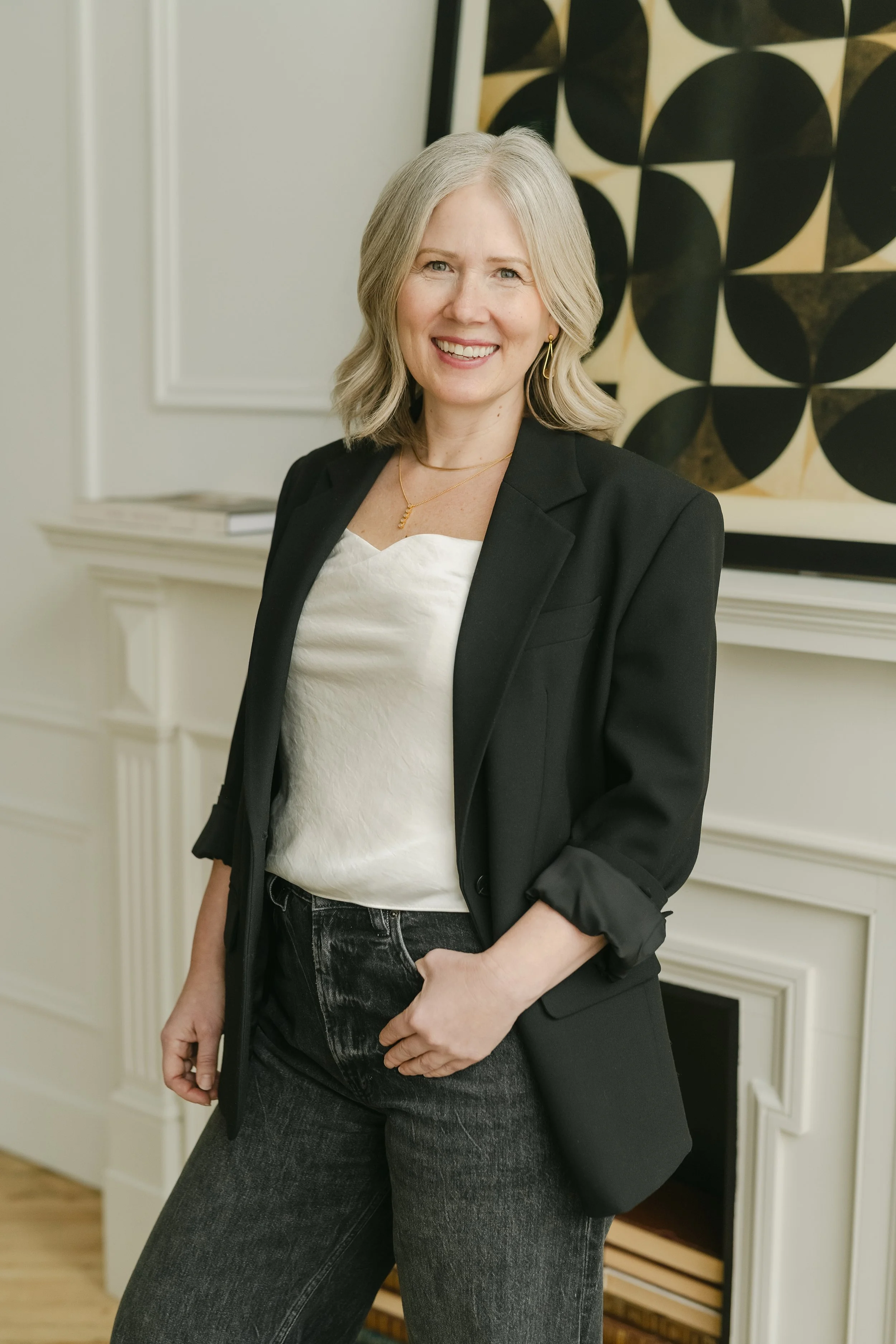 A woman with shoulder-length blonde hair wearing a blue pleated blouse and beige pants sitting on a cream-colored modern chair in a cozy living room with a fireplace, books, and a wooden floor.