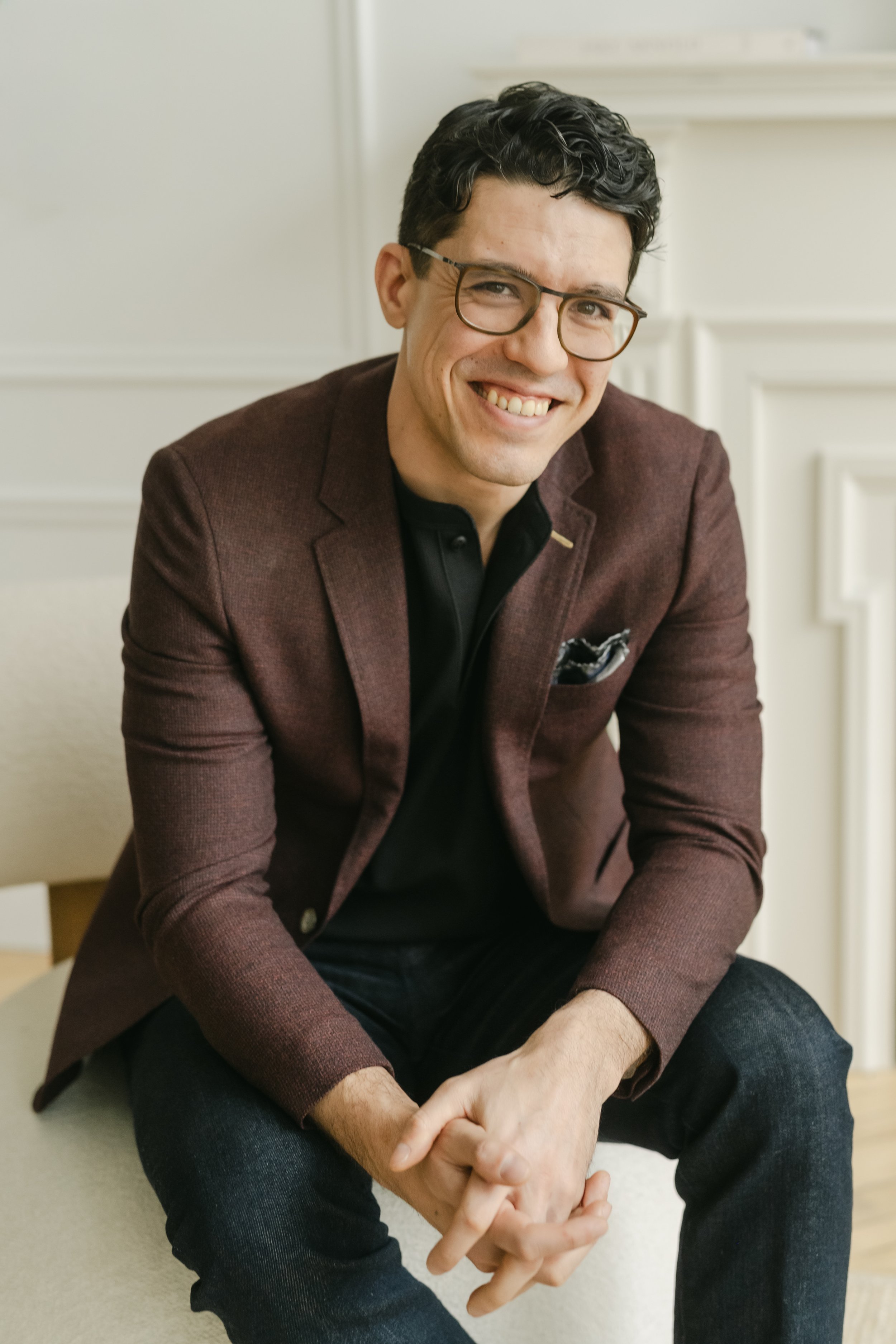 A man in a dark blazer, b;acl shirt, and black pants sits on a wooden stool, smiling, with a light-colored curtain in the background.