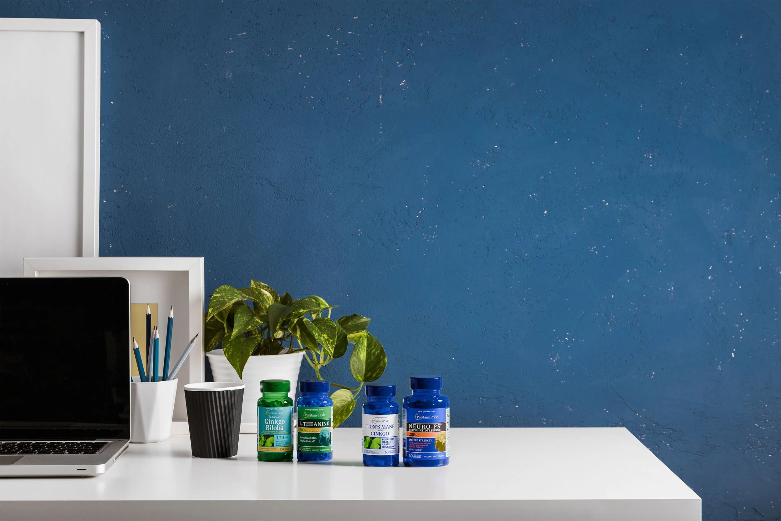 A white desk with a silver laptop, a white cup holding pencils, a potted green plant, and four bottles of supplements or vitamins against a blue textured wall.