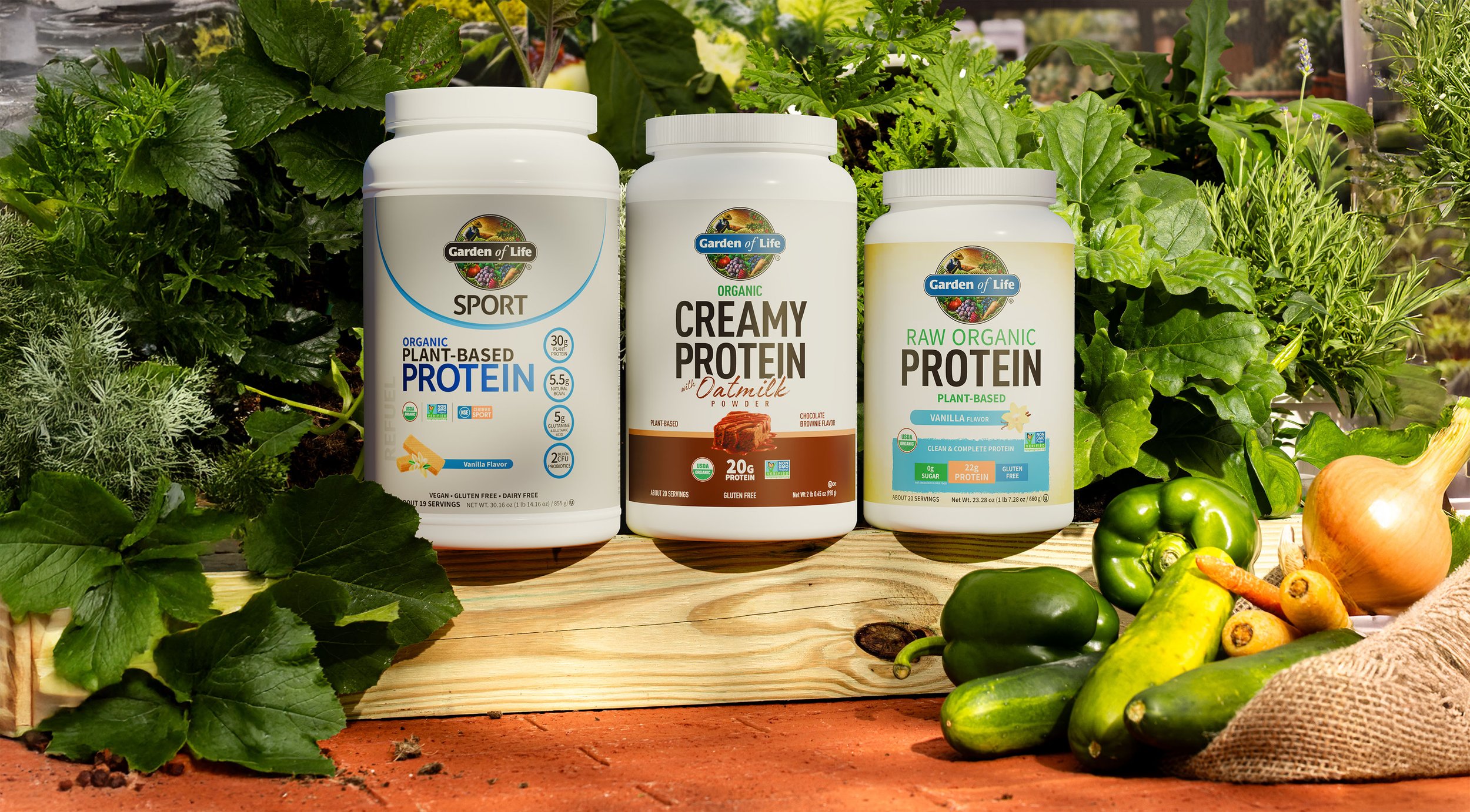 Three containers of plant-based protein supplements labeled 'Sport,' 'Creamy Protein,' and 'Raw Organic Protein' placed on a wooden surface outdoors, surrounded by fresh vegetables and green plants.