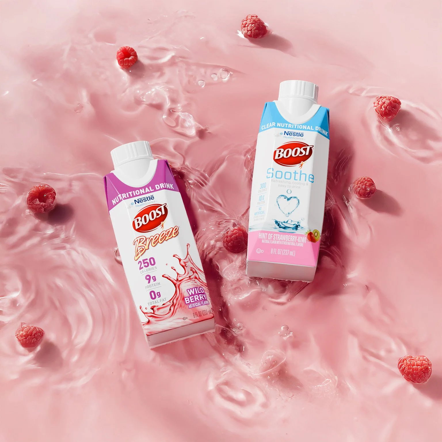 Two Boost nutritional drink cartons, one pink with wild berry flavor and one blue with strawberry kiwi flavor, placed on a swirling pink liquid surface with scattered raspberries.