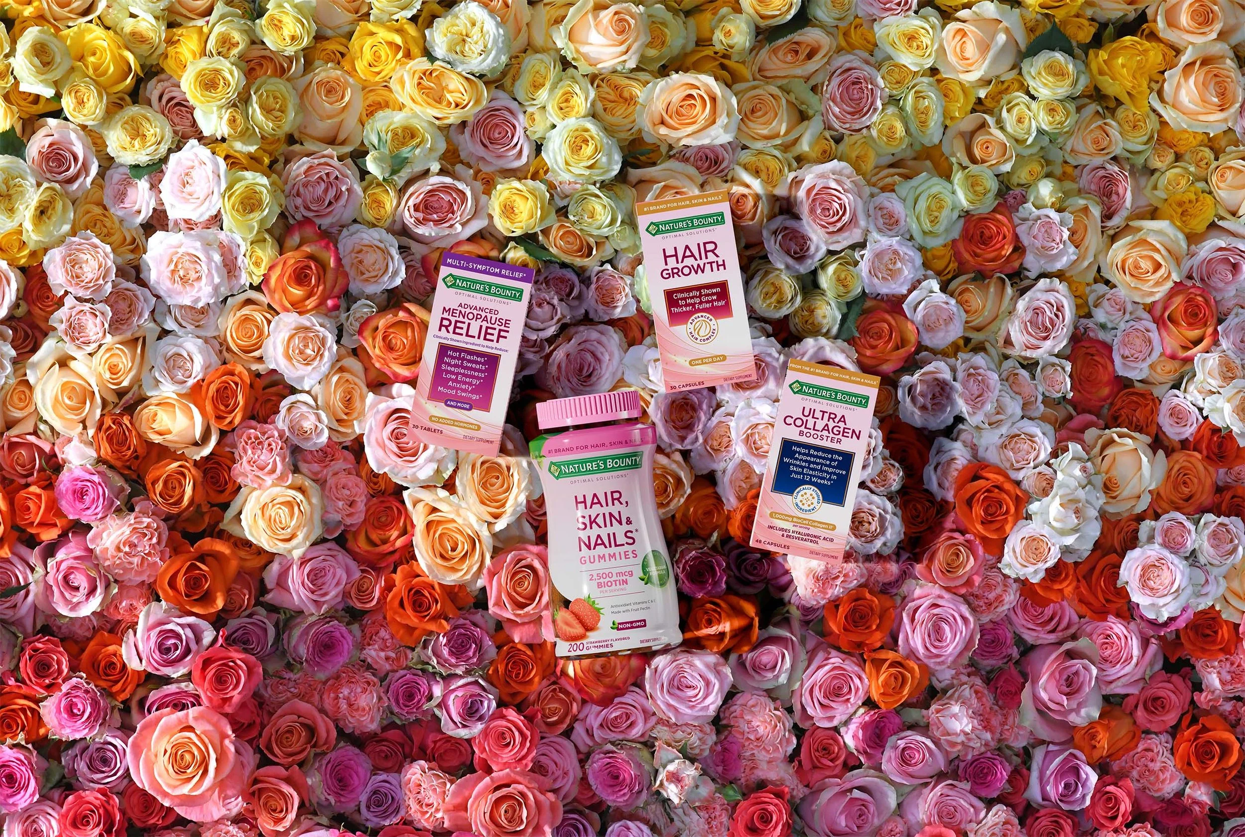 A large surface covered with a variety of colorful roses, including pink, white, orange, and peach. Four bottles of Nature's Bounty dietary supplements are placed on top of the roses, including hair growth, menopause relief, collagen booster, and gum