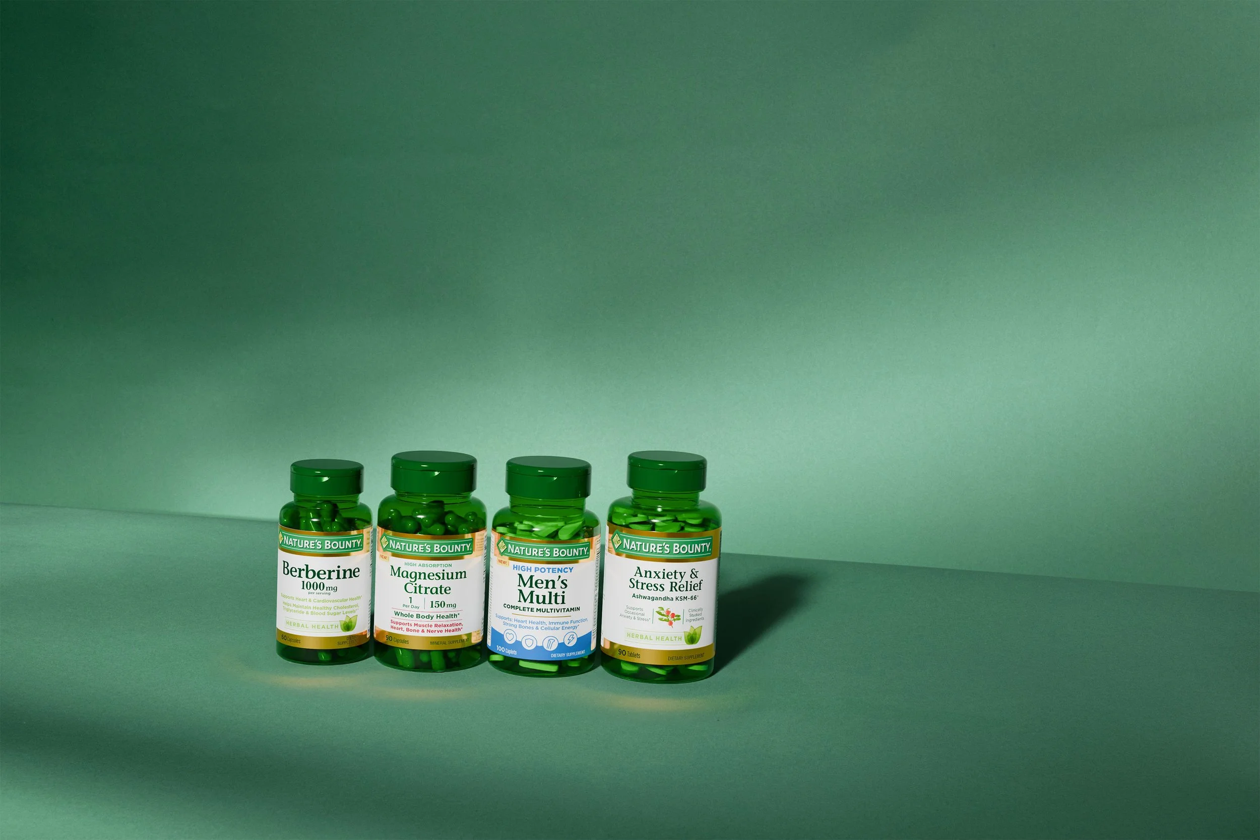 Four green bottles of Nature's Bounty supplements, including Berberine, Magnesium Citrate, Men's Multi, and Anxiety & Stress Relief, lined up on a green surface with a green gradient background.