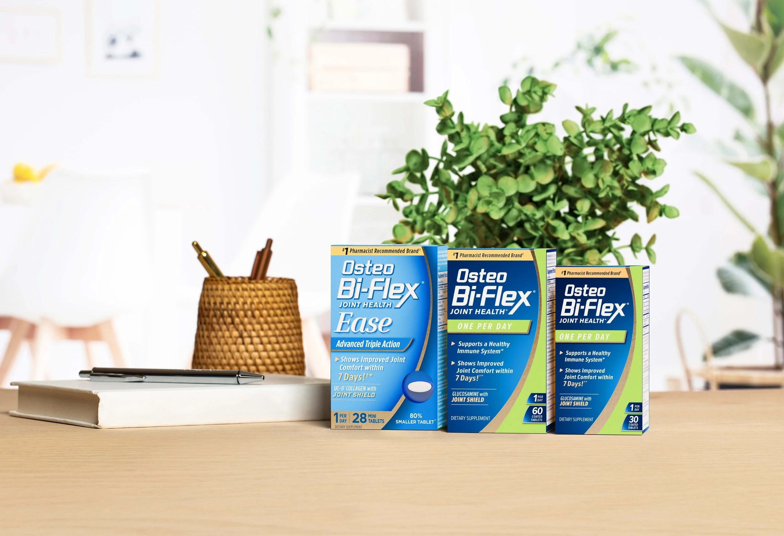 Three boxes of Osteo Bi-Flex Joint Health supplements on a light wooden table, with a pen and a basket holding pens or pencils, in a bright room with green plants and blurred background.