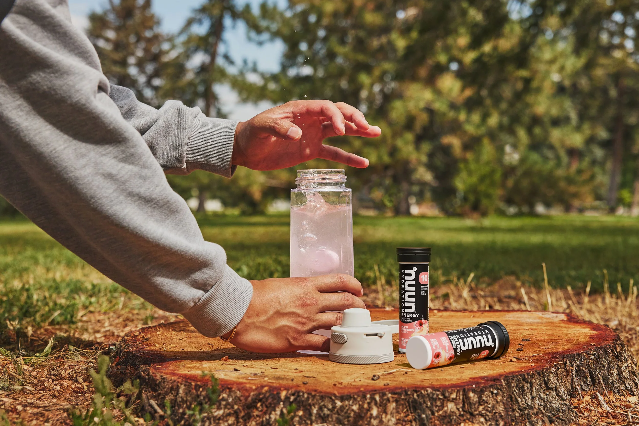 Person pouring water into a bottle outdoors on a tree stump, with supplement containers nearby.