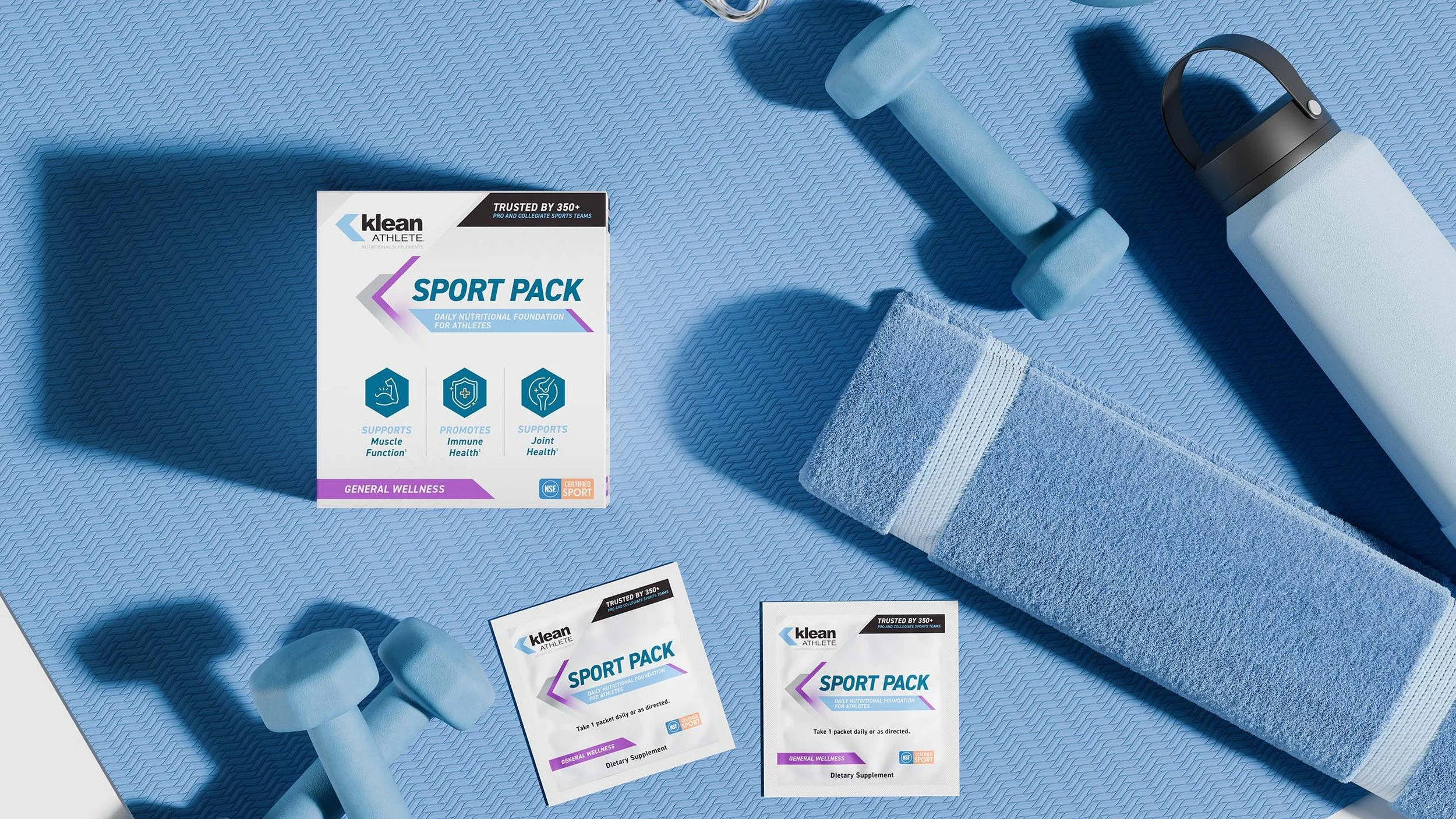 A blue textured workout mat with light blue dumbbells, a rolled-up towel, a water bottle, and packets of Klean Athlete Sport Pack dietary supplements.