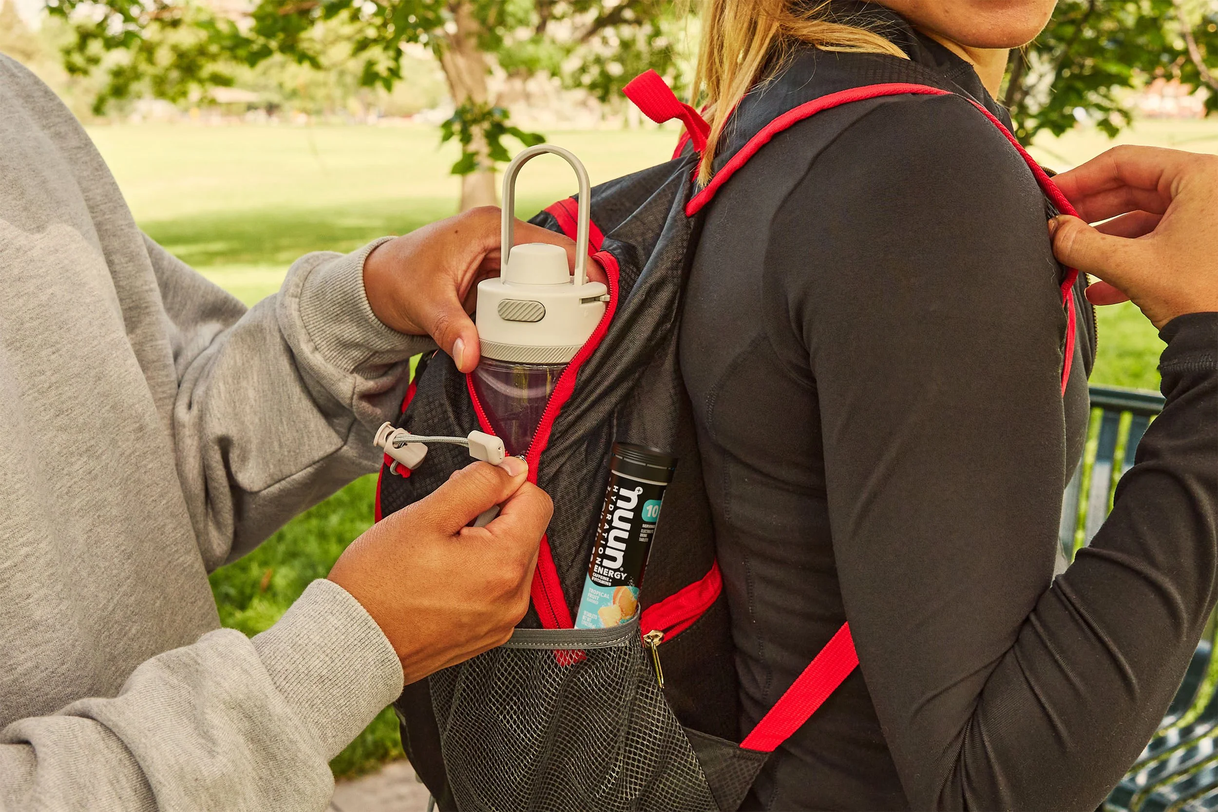 A person in a gray sweatshirt is placing a Nuun bottle into a pocket on the back of another person wearing a black jacket with red accents, who has a backpack.
