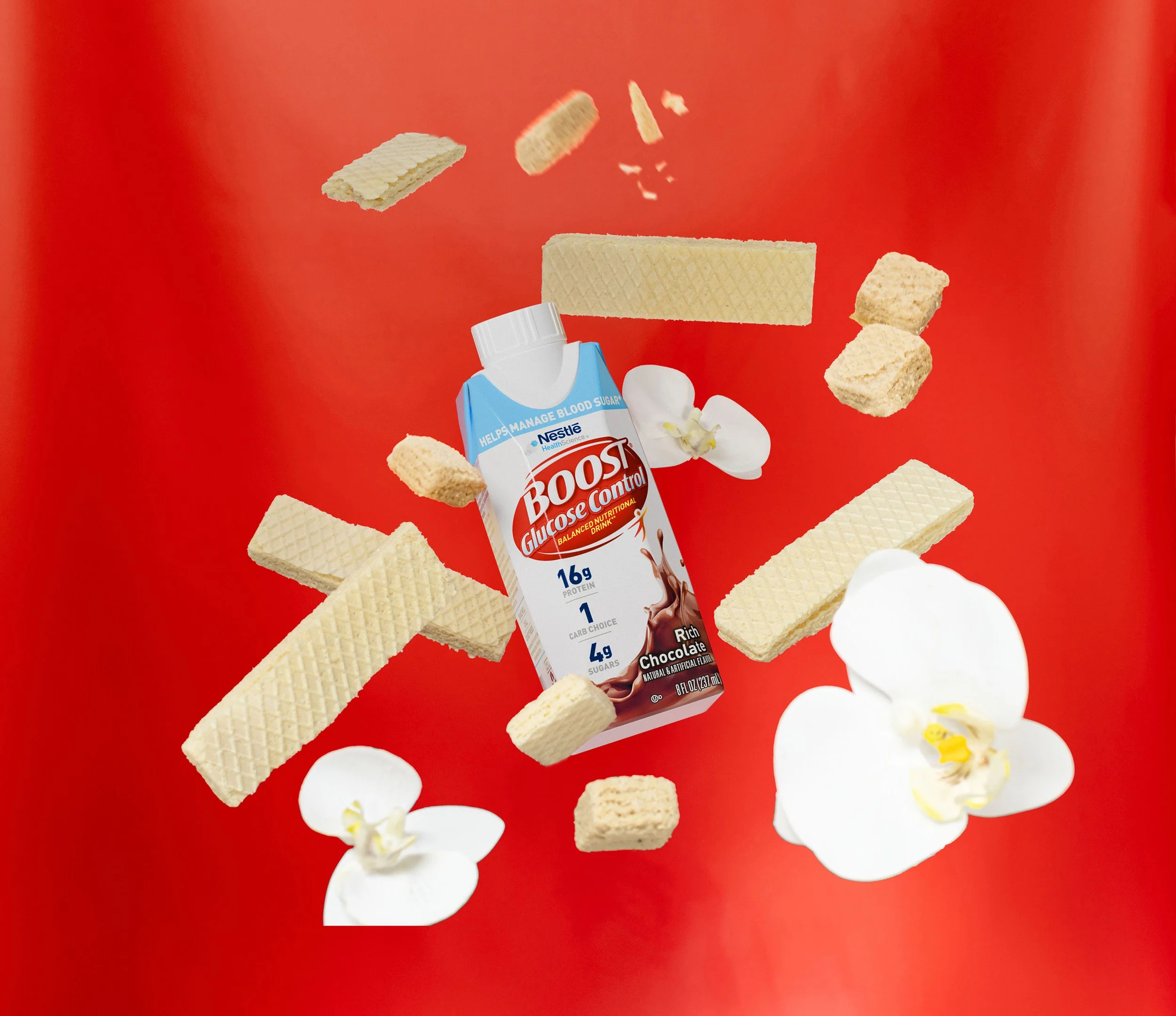 A bottle of Boost Glucose Control chocolate nutritional drink surrounded by wafer cookies, cereal pieces, and white flowers on a red background.