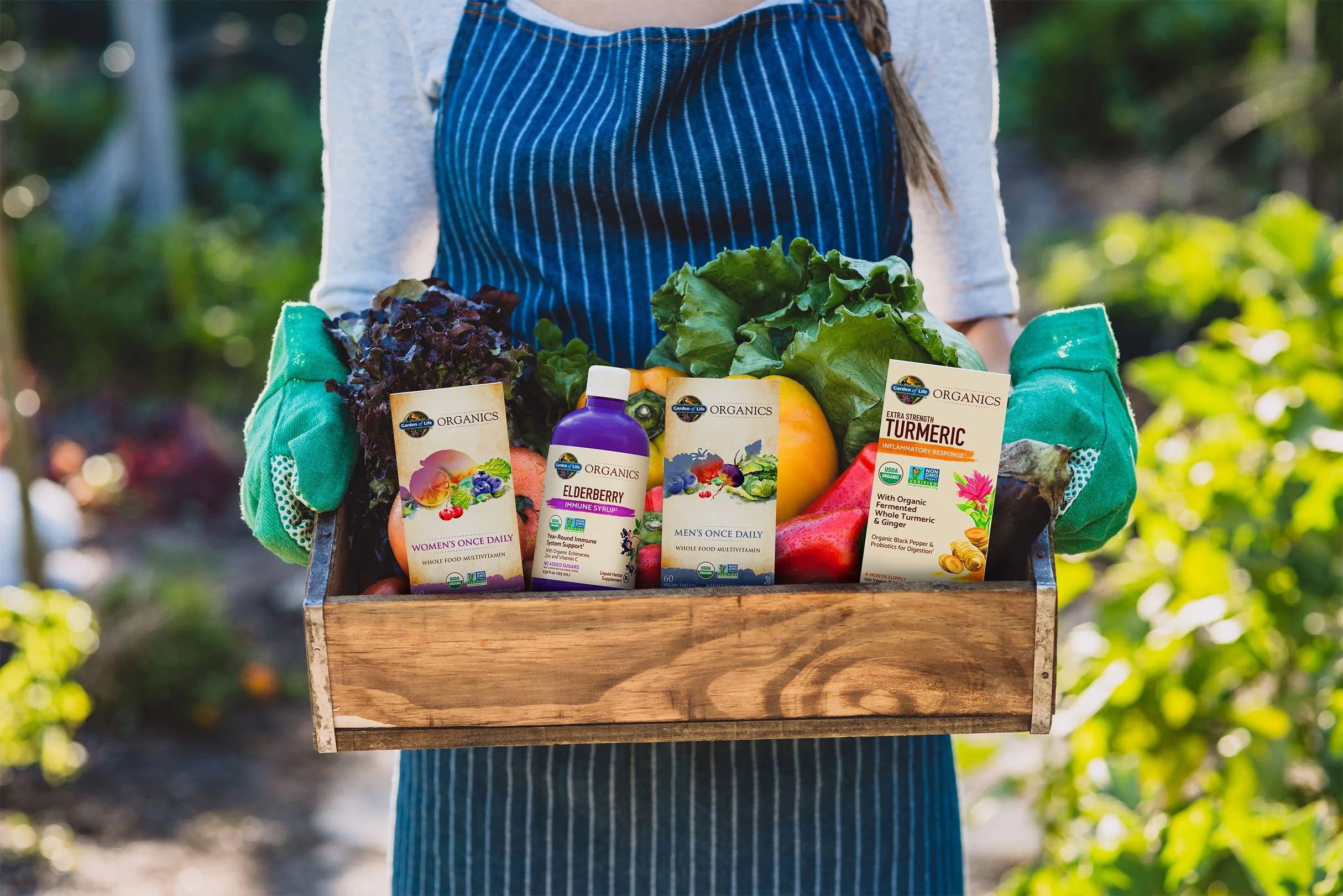 Person holding a wooden crate filled with fresh vegetables and health supplement bottles, outdoors in a garden.