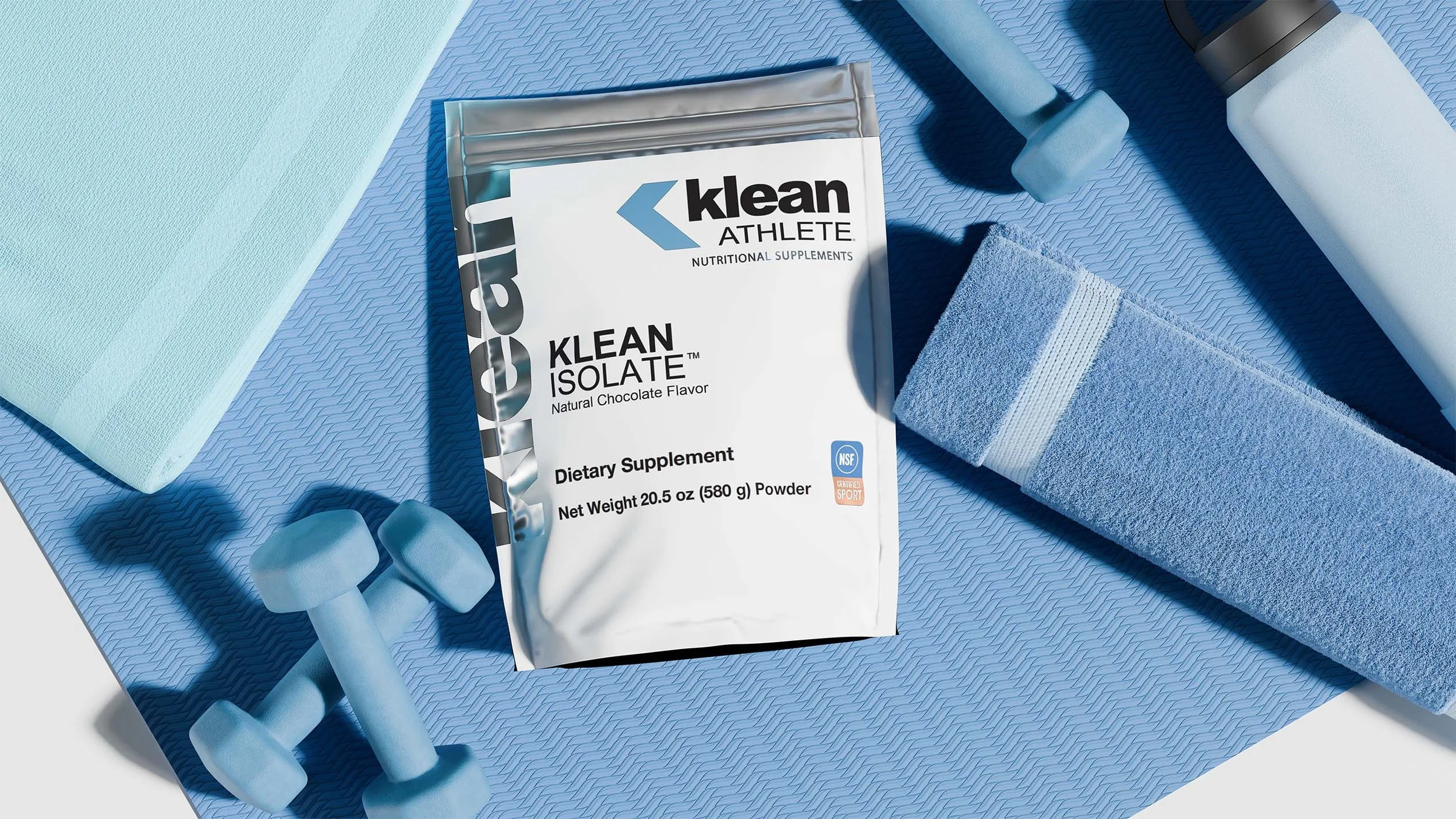 Workout supplies including a tube of Klean Isolate protein powder on a blue exercise mat, blue dumbbells, a rolled-up blue towel, and two foam rollers.