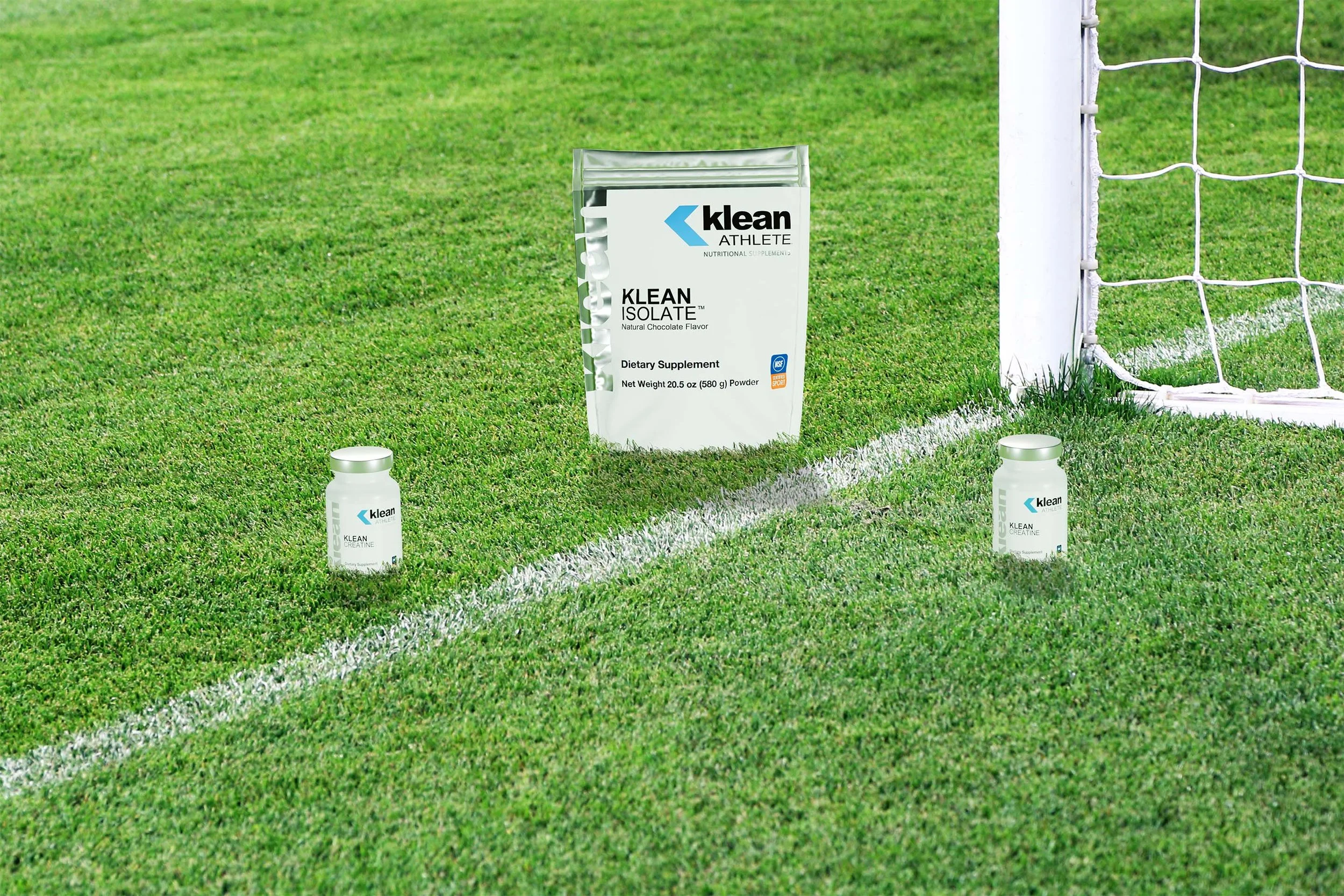Supplements on a soccer field near a goal post, including a bag labeled 'Klean Isolate,' and two bottles labeled 'Klean Creatine.'