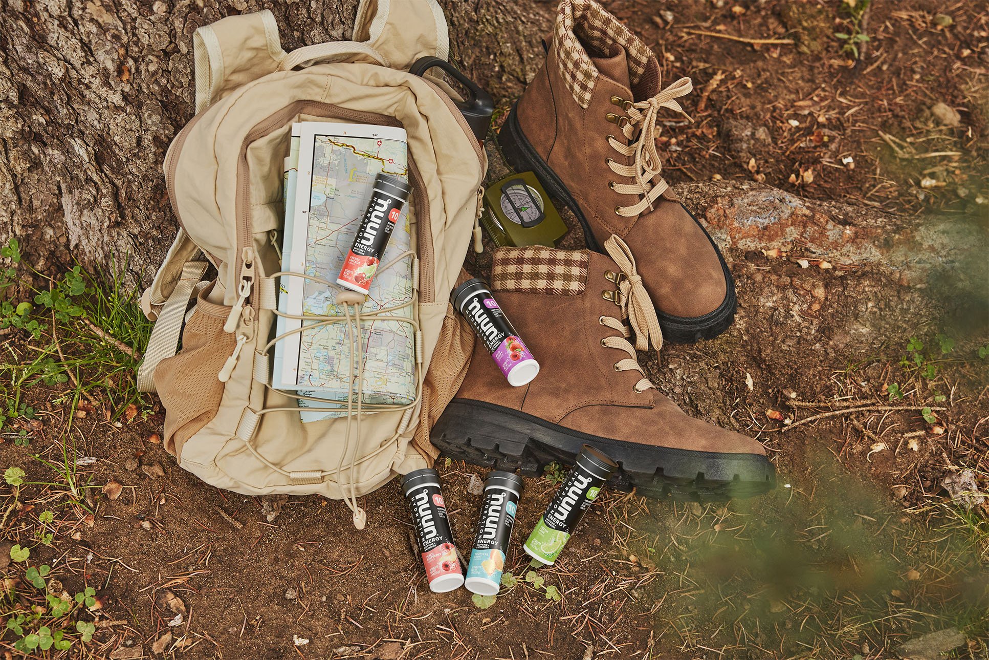 A pair of brown hiking boots, a beige backpack with a map and energy drink bottles, a small green compass, and a portable black radio on the ground next to a tree.