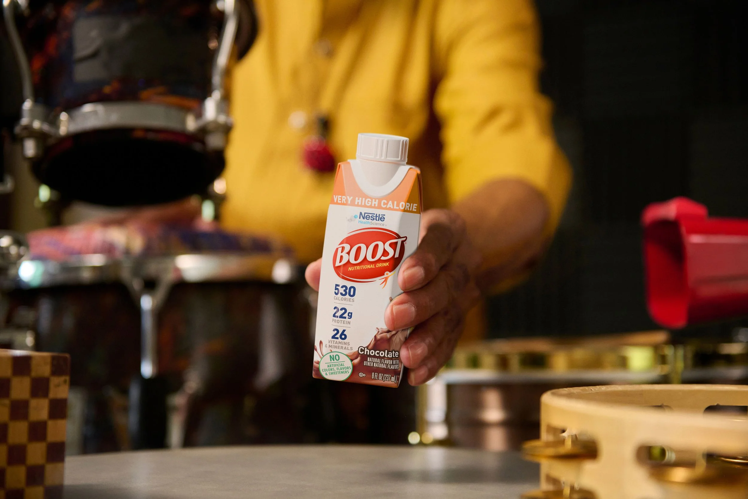 Person wearing a yellow shirt holding a Boost nutritional drink carton in a kitchen or food preparation area.