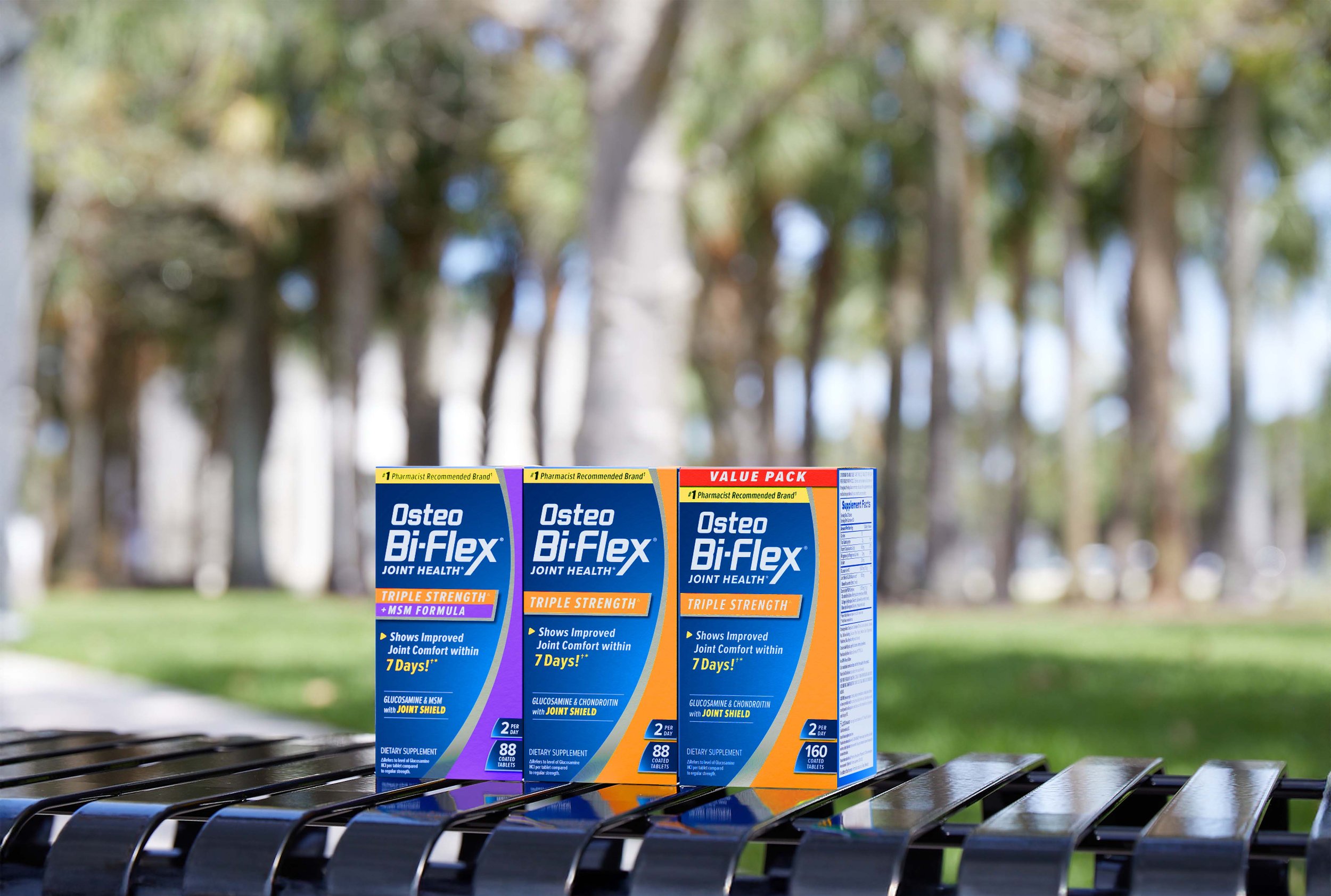 Three boxes of Osteo Bi-Flex joint health supplement on a park bench, with a background of trees and grass.