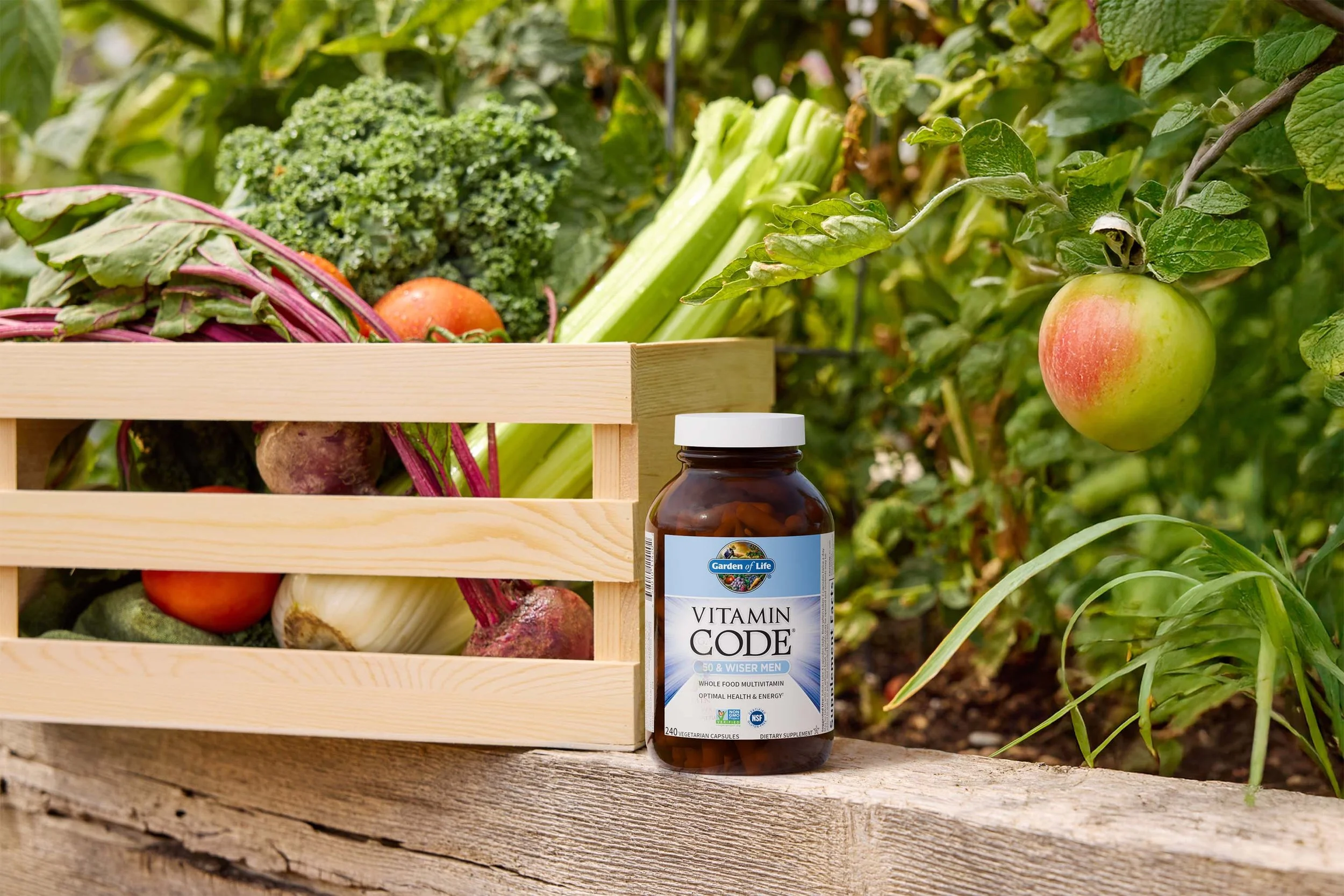 A bottle of Garden of Life Vitamin Code multivitamins placed on a wooden surface next to a wooden crate filled with fresh vegetables, set outdoors on a garden table with green plants and an apple tree in the background.
