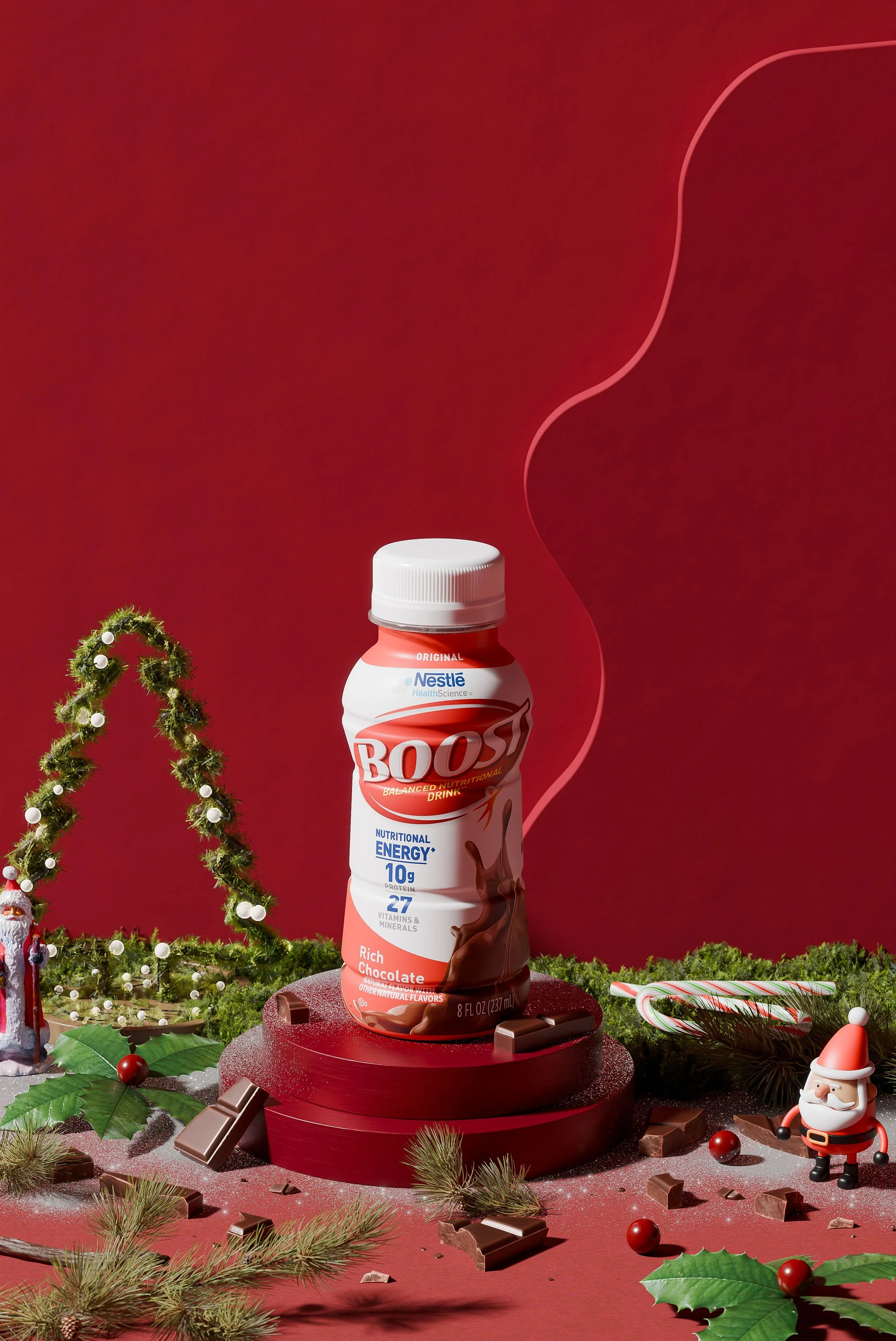 A bottled Boost chocolate energy drink on a red pedestal with Christmas decorations including a Santa figurine, holly leaves, pine branches, candy canes, and scattered chocolate pieces on a red background.