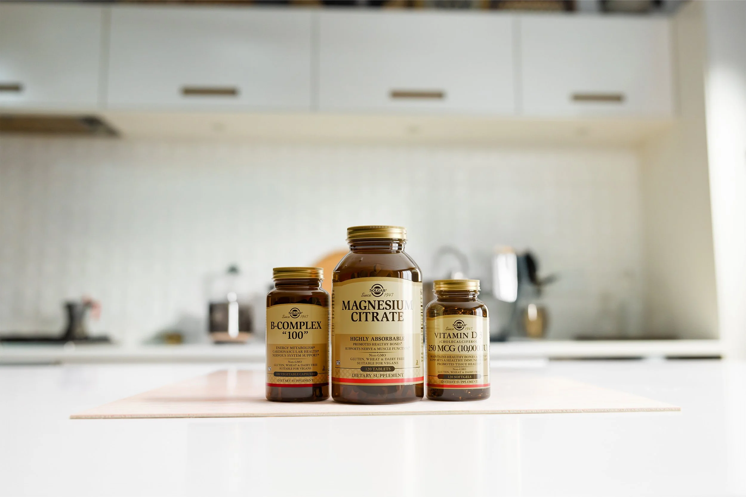 Three brown supplement bottles labeled magnesium citrate, B-complex, and vitamin D, placed on a white countertop in a kitchen.