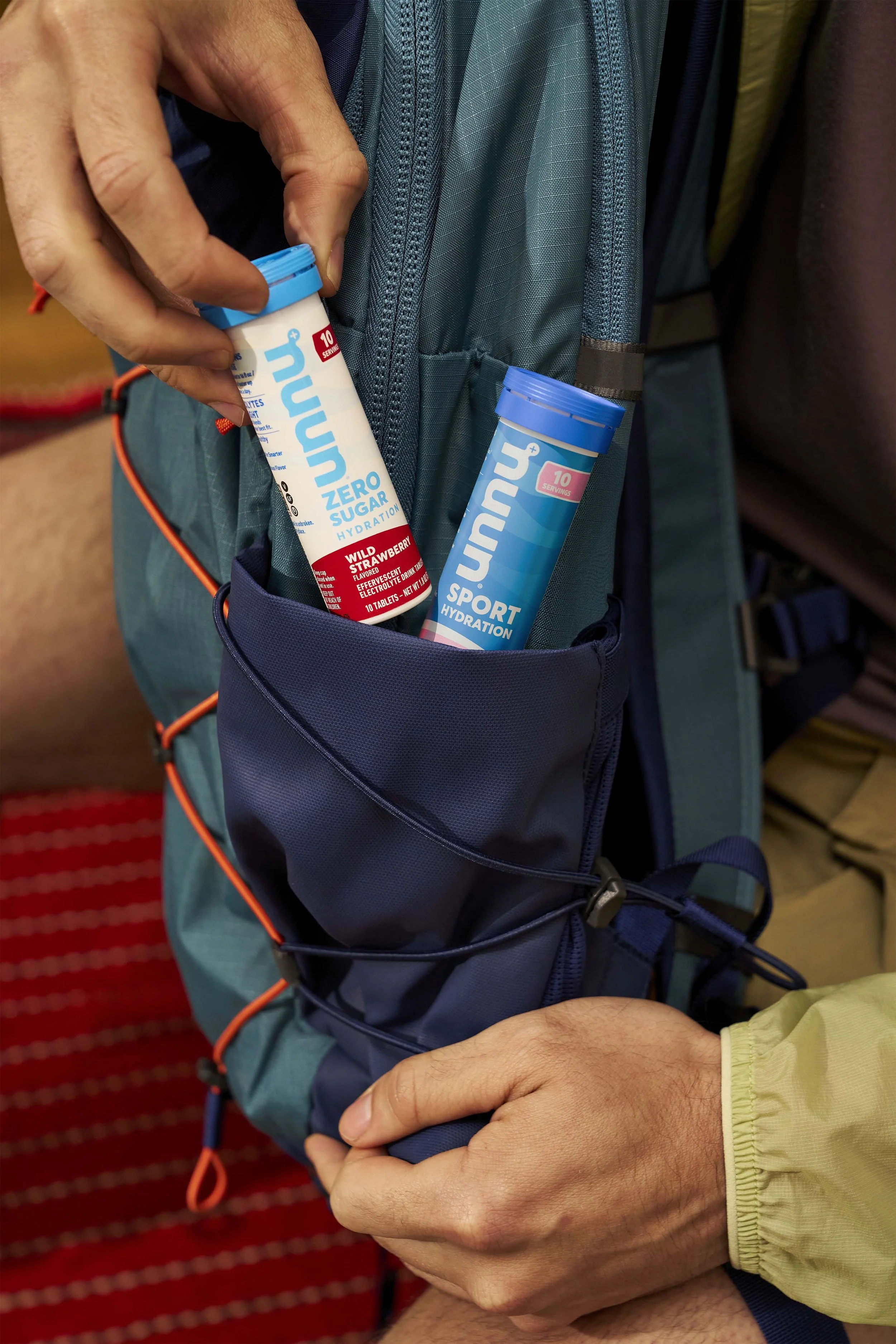 Person placing two tubes of Nuun hydration tablets, one wild strawberry flavor and the other sport hydration, into the side pocket of a blue backpack.