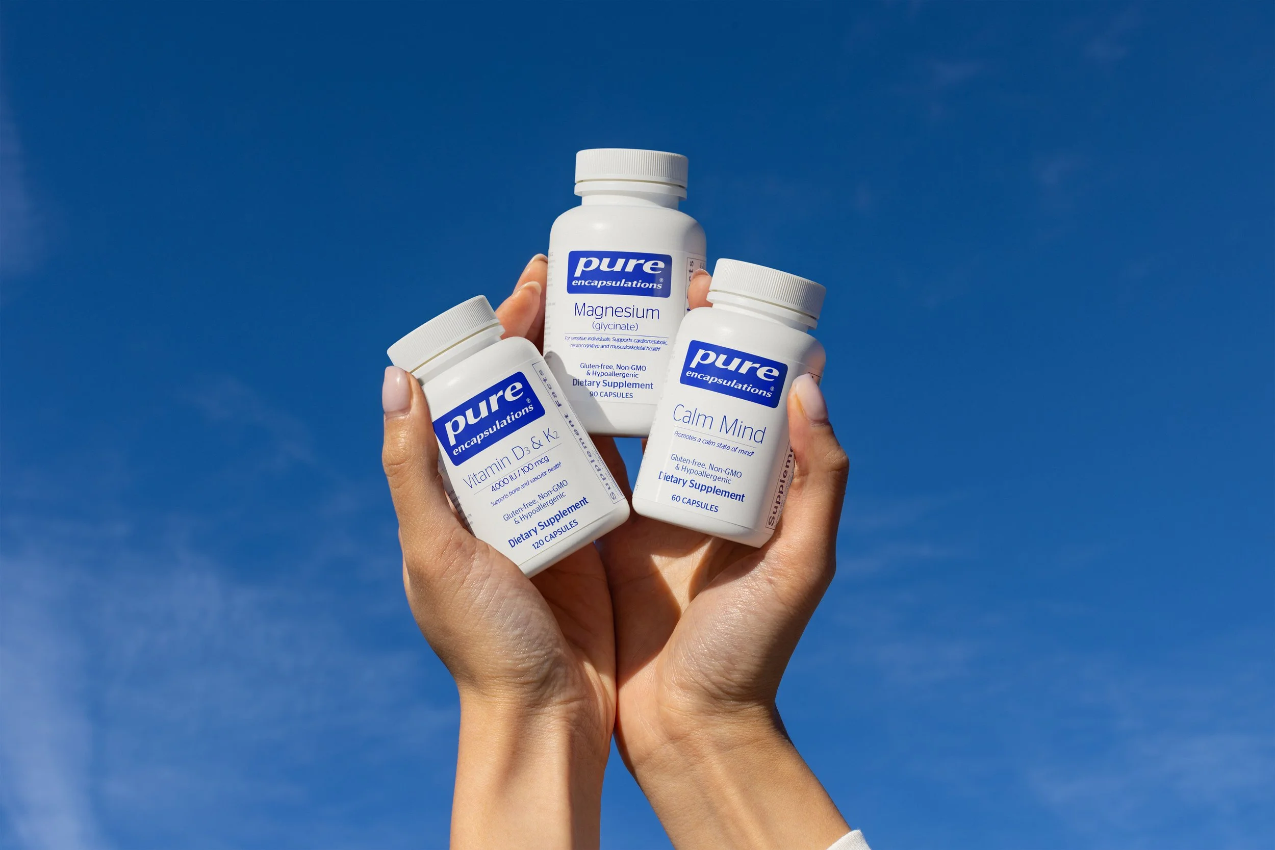 Person holding three bottles of dietary supplements against a clear blue sky, labeled for magnesium, vitamin D & K2, and calming mind.