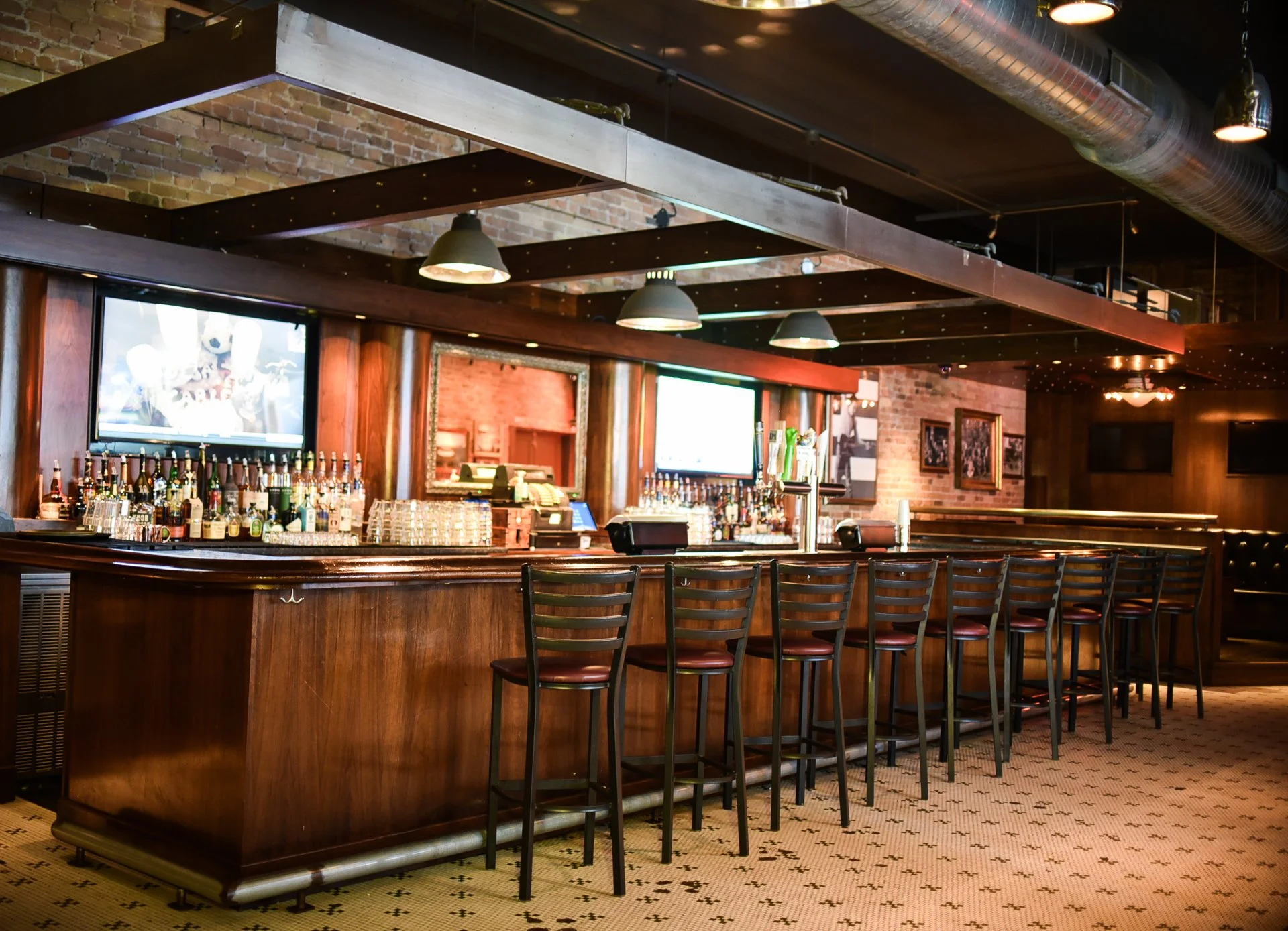 Bar counter with bottles of alcohol, bar stools, and two television screens in a dimly lit restaurant or pub with exposed brick walls and wooden accents.