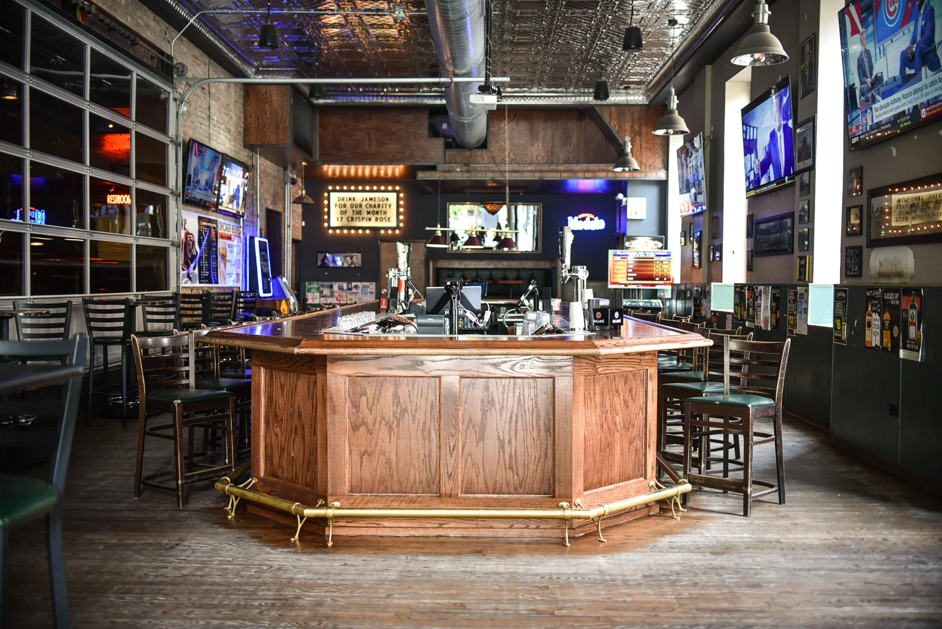 Empty sports bar with a central wooden bar counter surrounded by chairs and barstools, multiple TV screens on the walls, and neon signs.