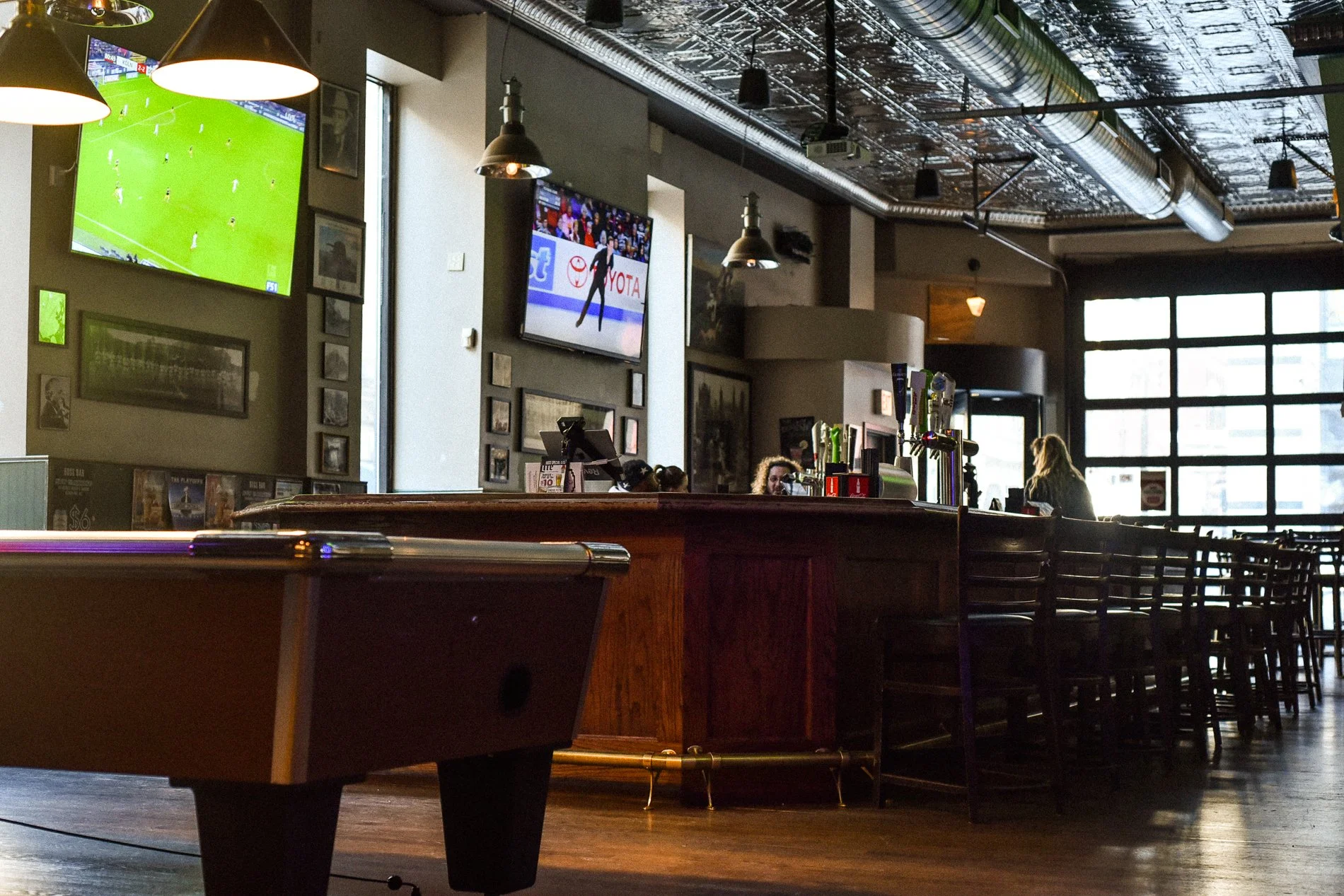 Interior of a sports bar or pub with a pool table, bar counter, and seating area. Multiple televisions display a soccer game and a figure skater. Several hanging lights and a large window provide natural light.
