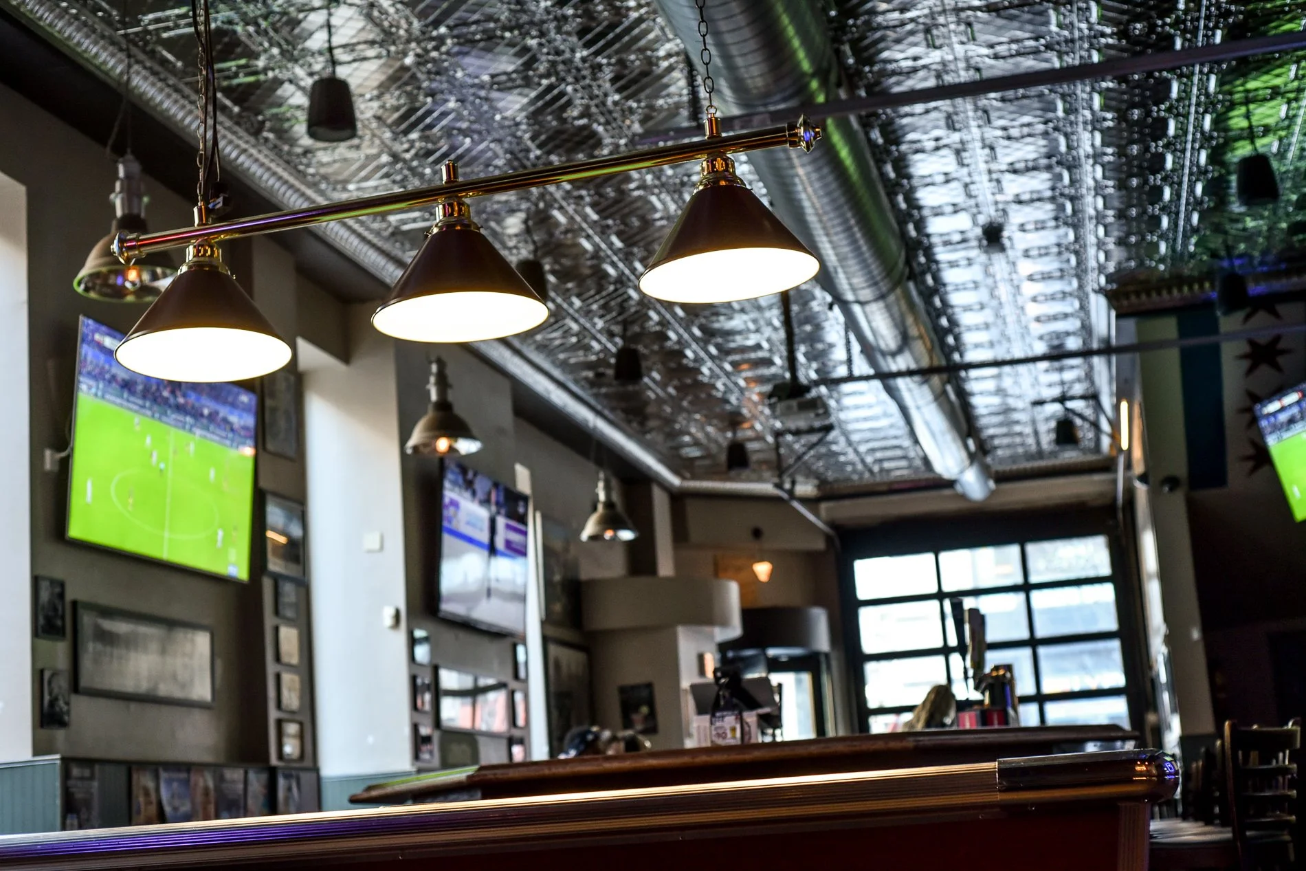 Interior of a bar or restaurant with hanging ceiling lights, multiple TV screens showing a sports event, and decorative framed pictures on the wall. Large windows in the background let in natural light.