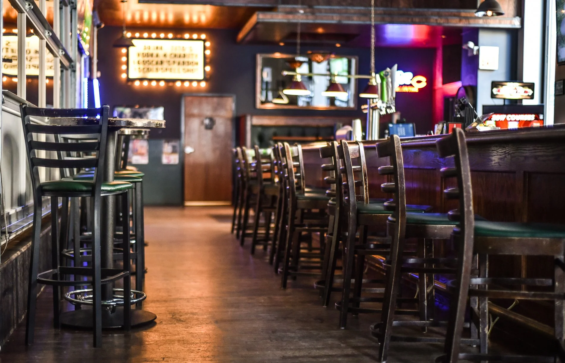 Empty bar with wooden counter and chairs, neon signs, and a illuminated menu board in a dimly lit pub or bar.