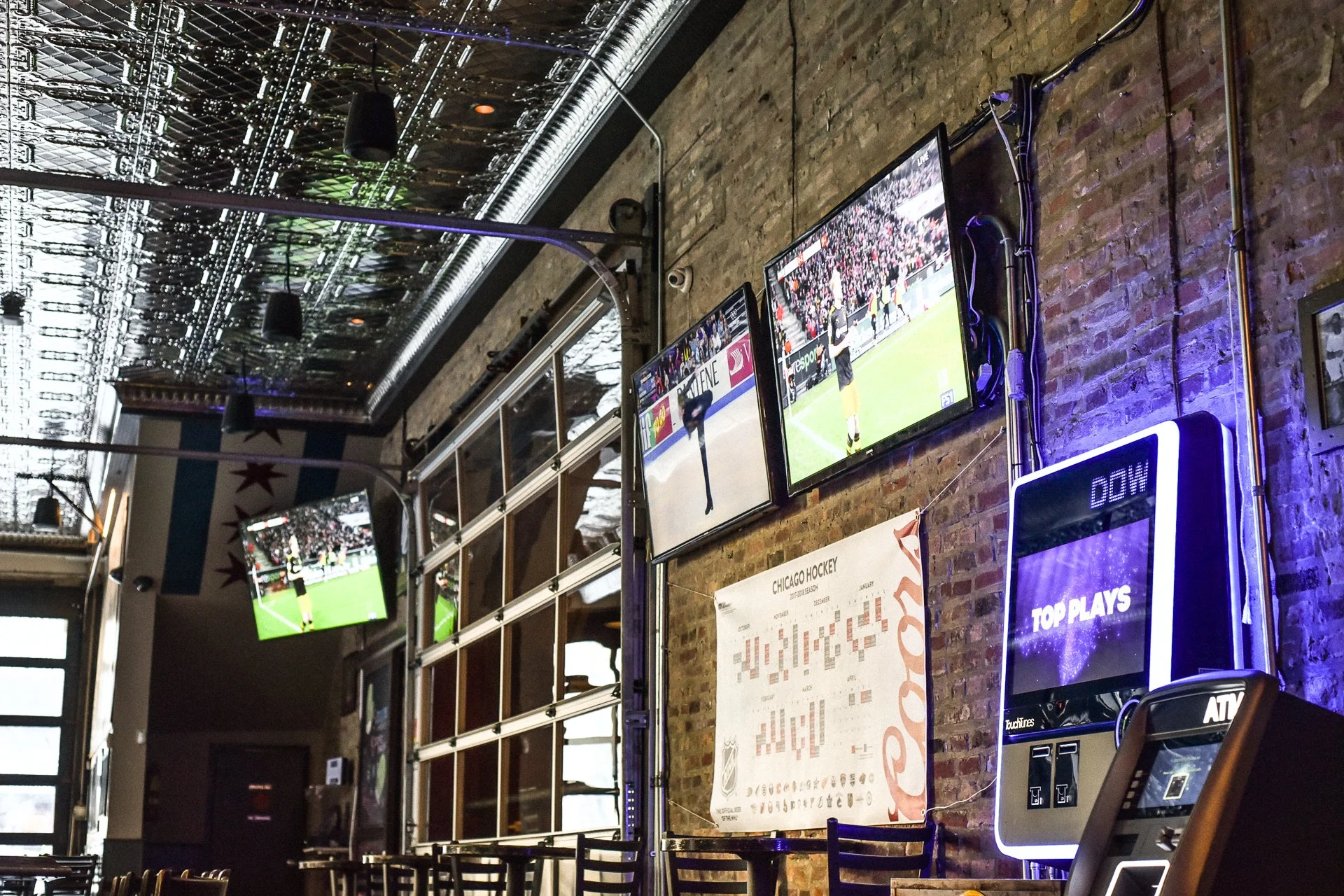 Several television screens displaying a soccer game, mounted on an exposed brick wall with visible wires. There is a Chicago Hockey poster on the wall and a digital scoreboard showing 'TOP PLAYS' and 'DOWN'.