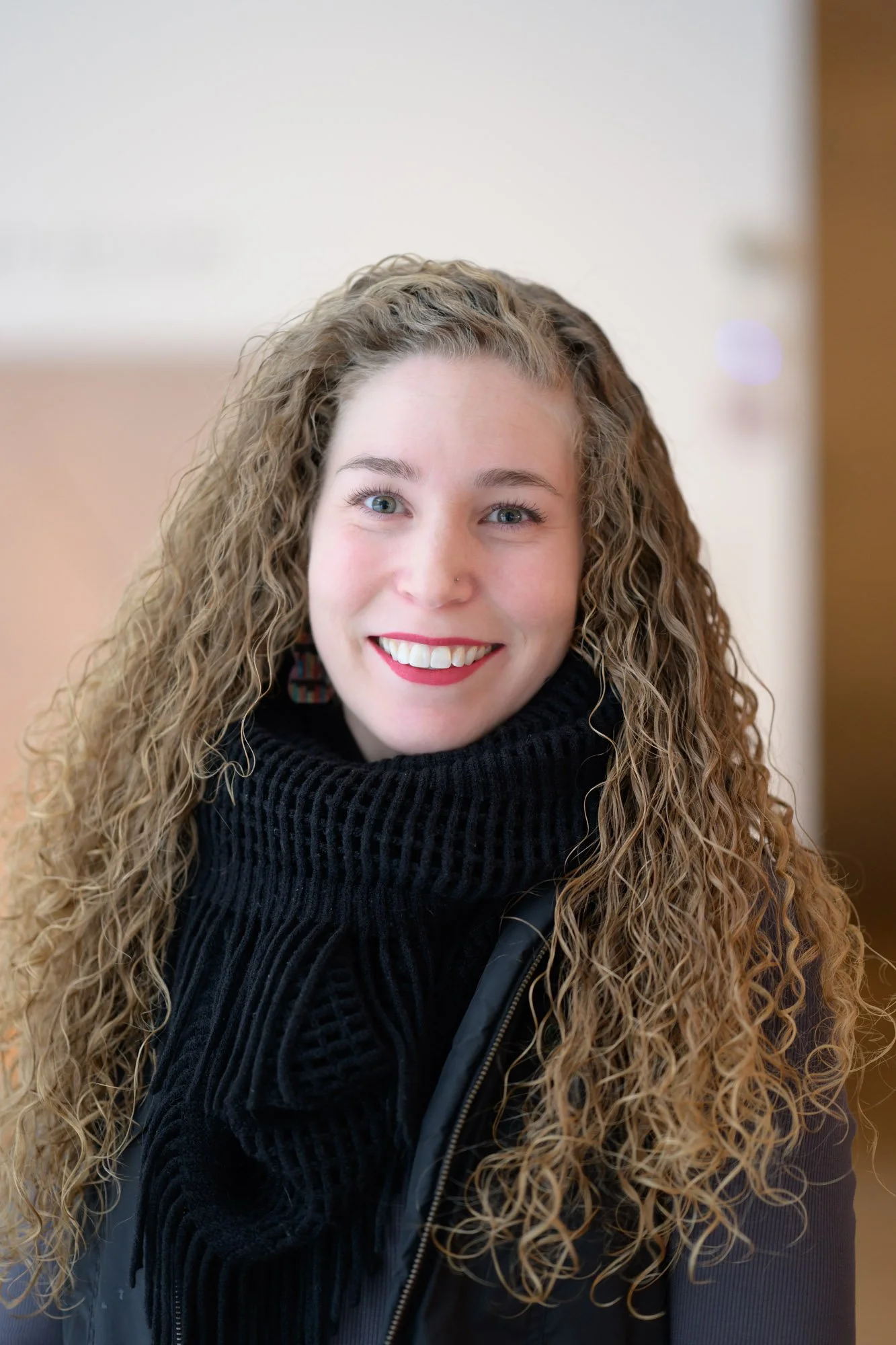 Portrait of a woman with curly blonde hair, wearing a black blazer and a patterned scarf, standing in front of an abstract colorful background, smiling with crossed arms.