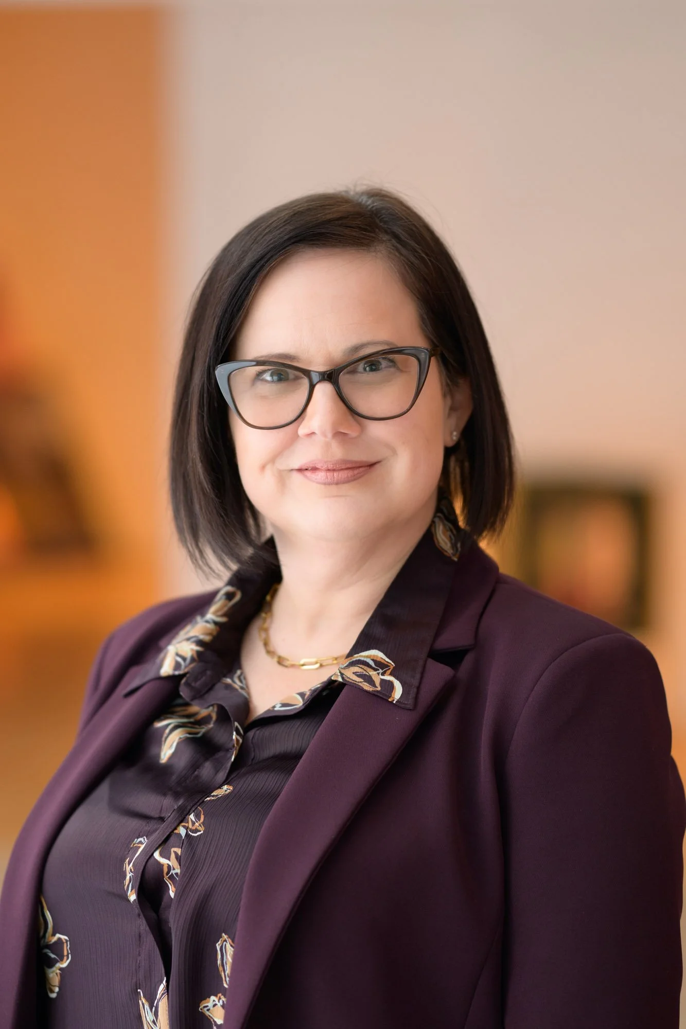 A woman with short black hair, wearing a floral-patterned brown and black blouse and gold earrings, posing in front of a colorful abstract background.