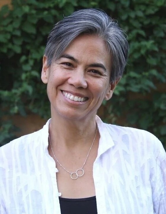 A smiling woman with short, gray hair, wearing a white shirt and a necklace with two interlocking circles, standing outdoors with green foliage in the background.
