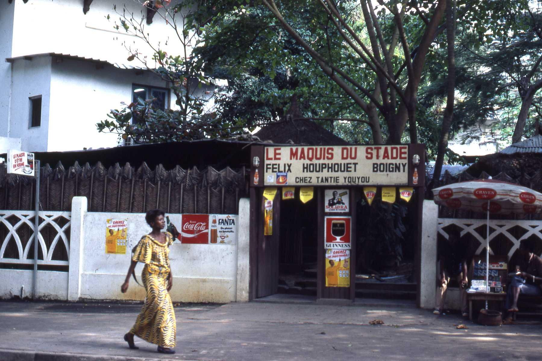 Woman in a yellow patterned dress walking past the entrance of Le Maquis du Stade Félix Houphouët-Boigny in Côte d’Ivoire