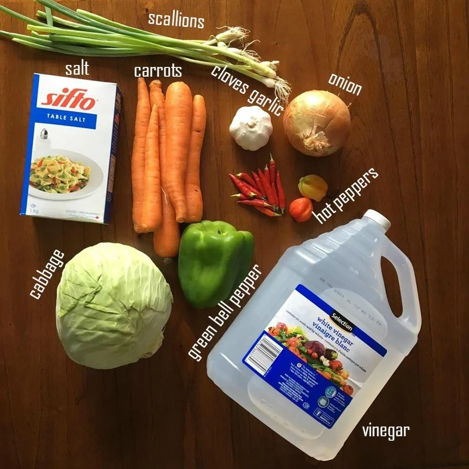 Labelled Haitian pikliz ingredients arranged on a wooden table, including cabbage, carrots, peppers, onion, scallions, garlic, salt, and vinegar