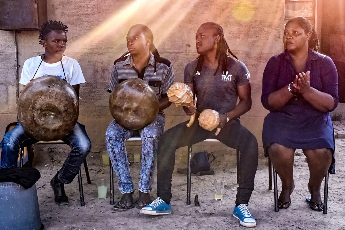 Four Zimbabwean musicians playing mbira and hosho while singing and clapping outside a home in Chitungwiza