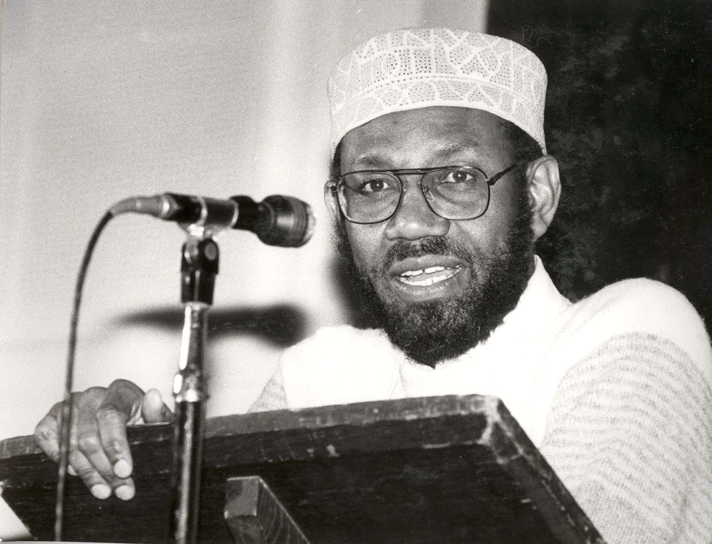 Black-and-white photograph of Abdilatif Abdalla wearing glasses and a patterned cap at a lectern beside a microphone