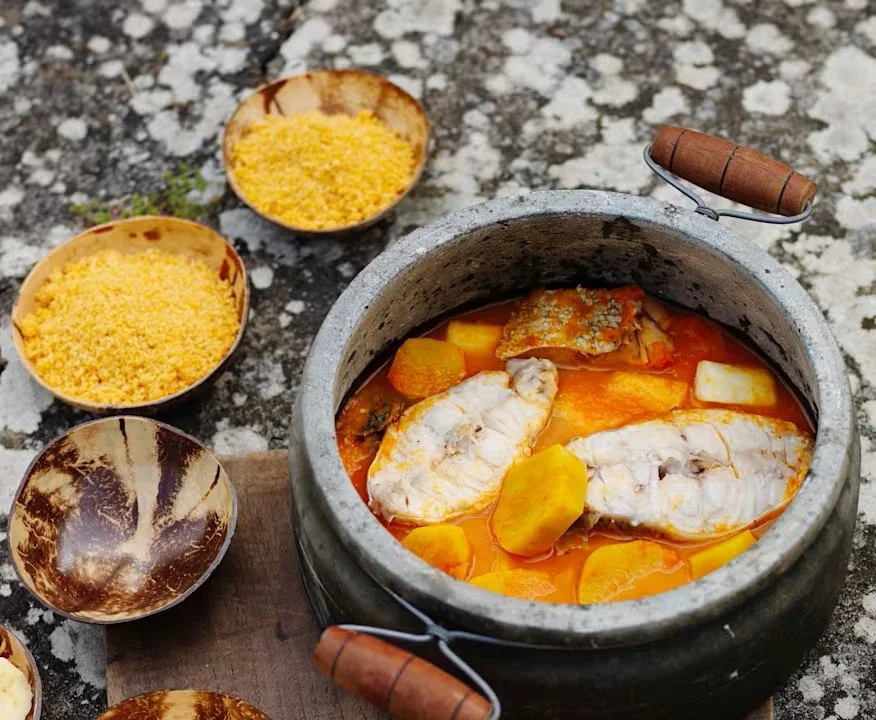 Pot of muzongué with fish steaks and sliced vegetables in orange broth, beside bowls of cassava flour.