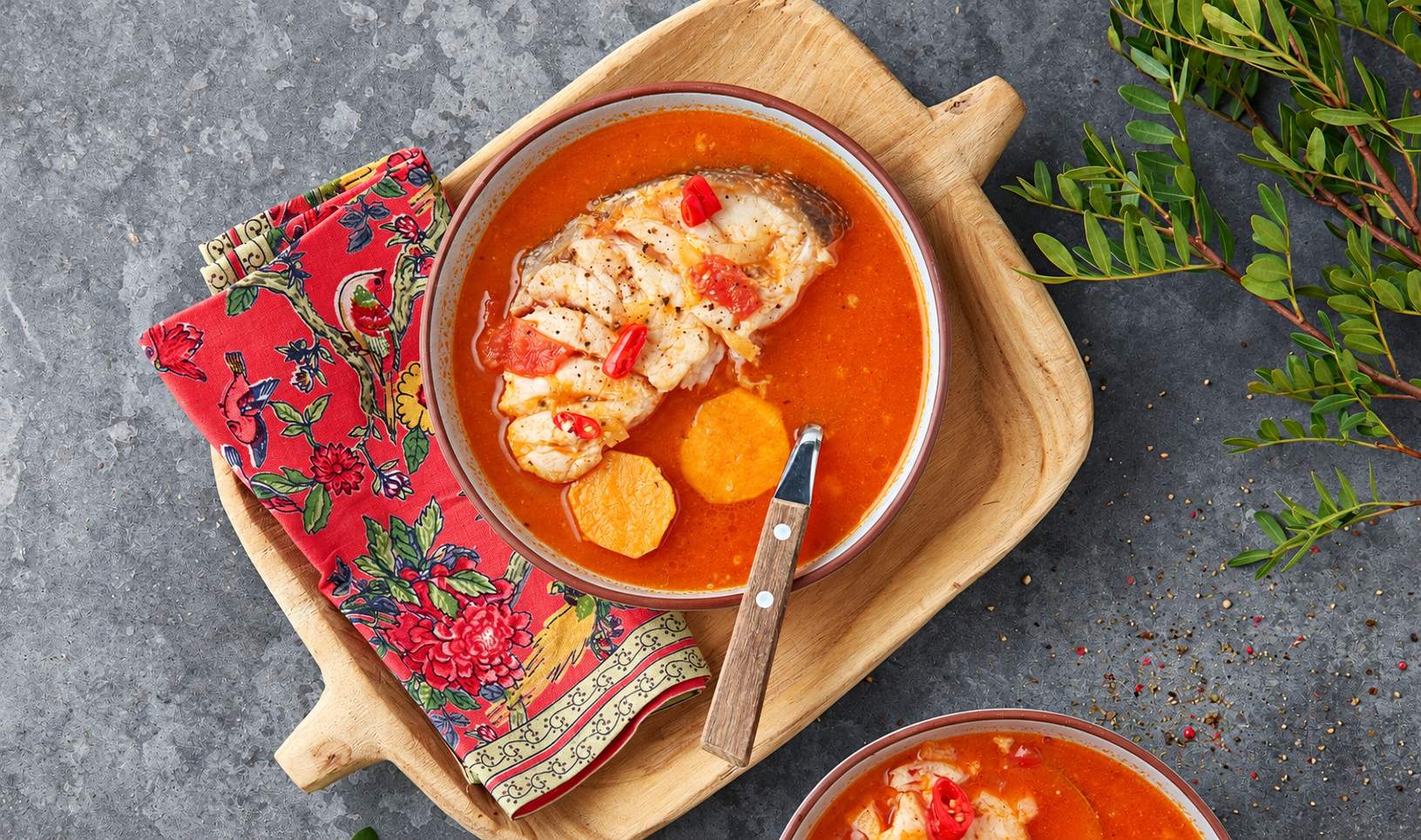 Overhead view of muzongué with fish, sweet potato rounds, and red chilli in a bowl on a wooden tray.
