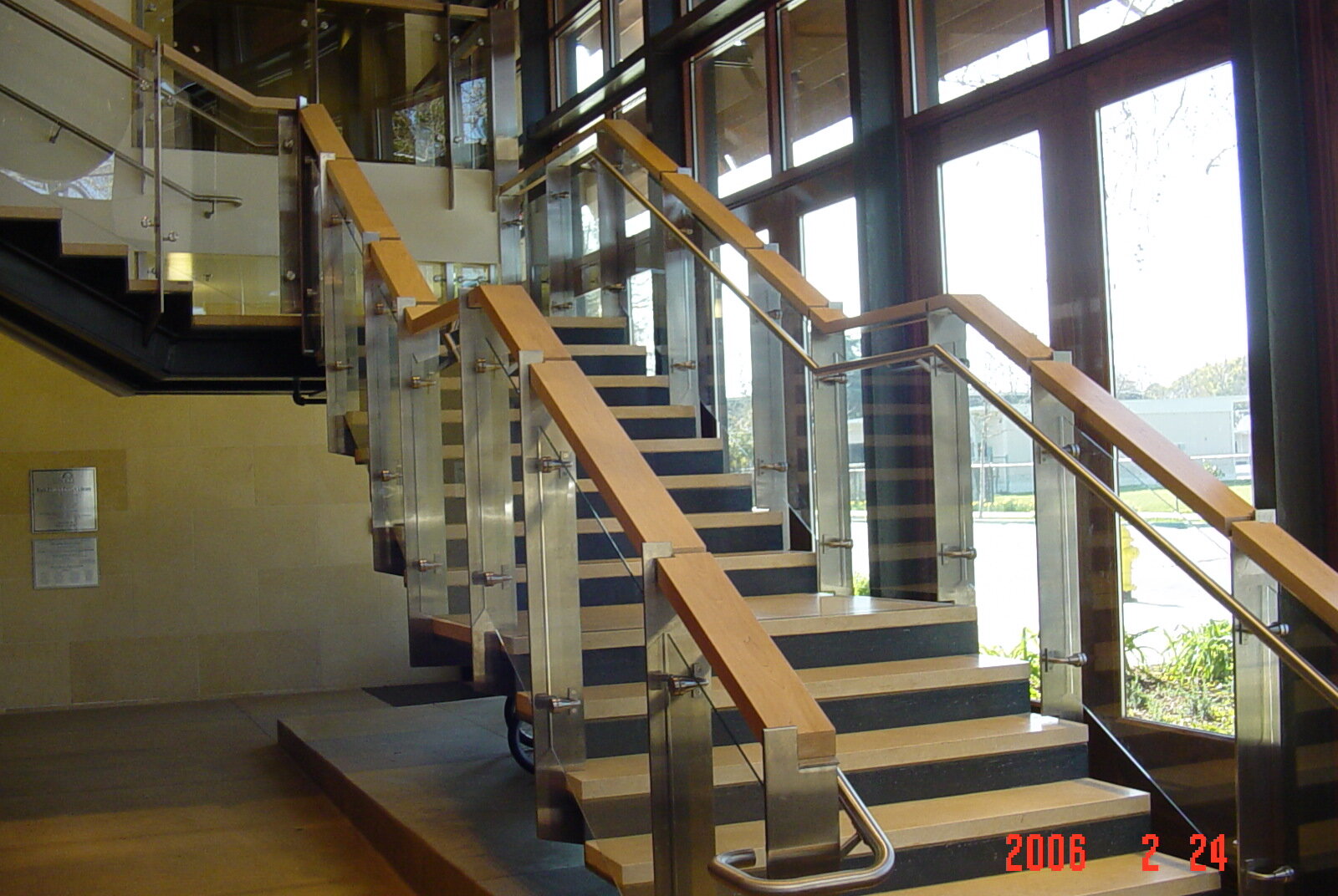 Interior view of a modern staircase with metal railings and wooden handrails, next to large glass windows letting in natural light.