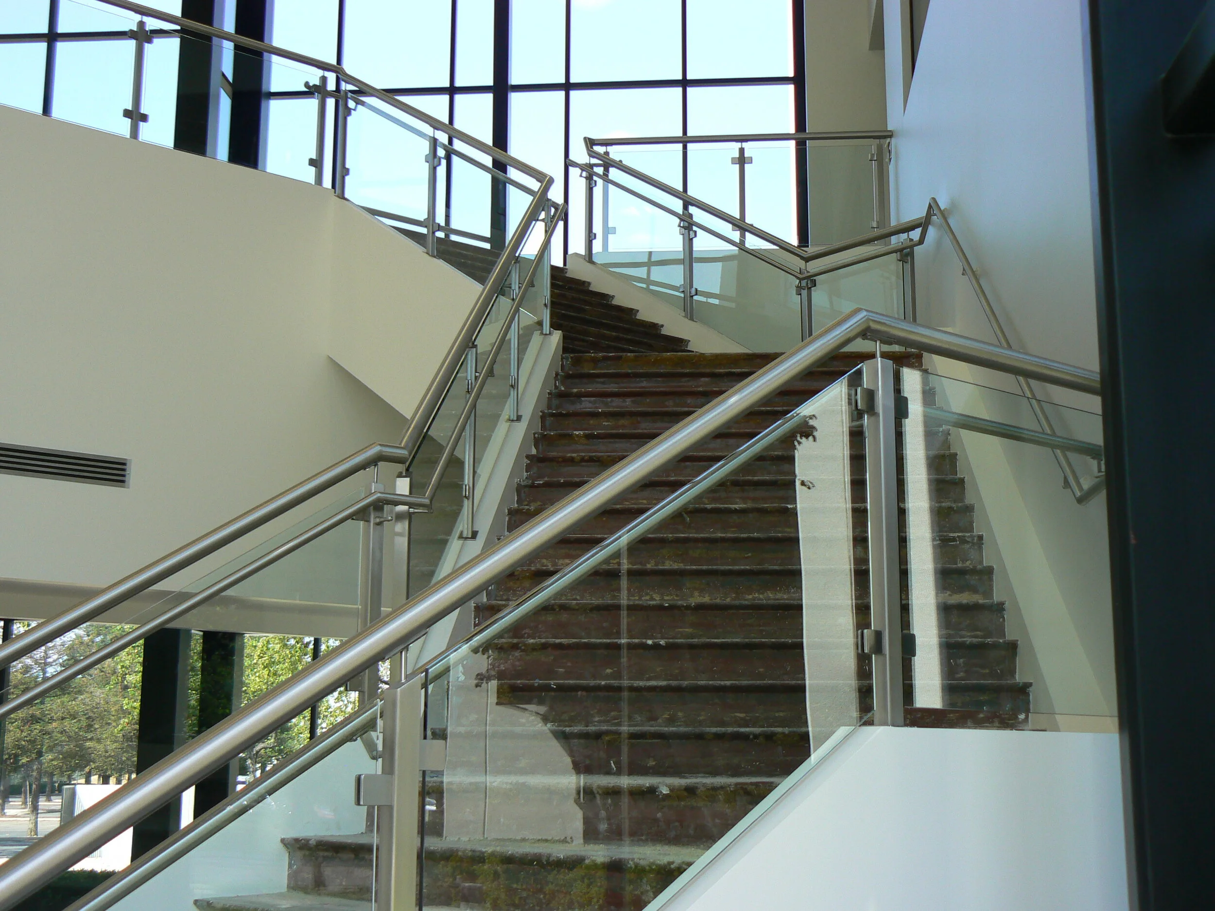 Indoor staircase with glass and metal railing, leading to an upper floor with large windows letting in natural light.