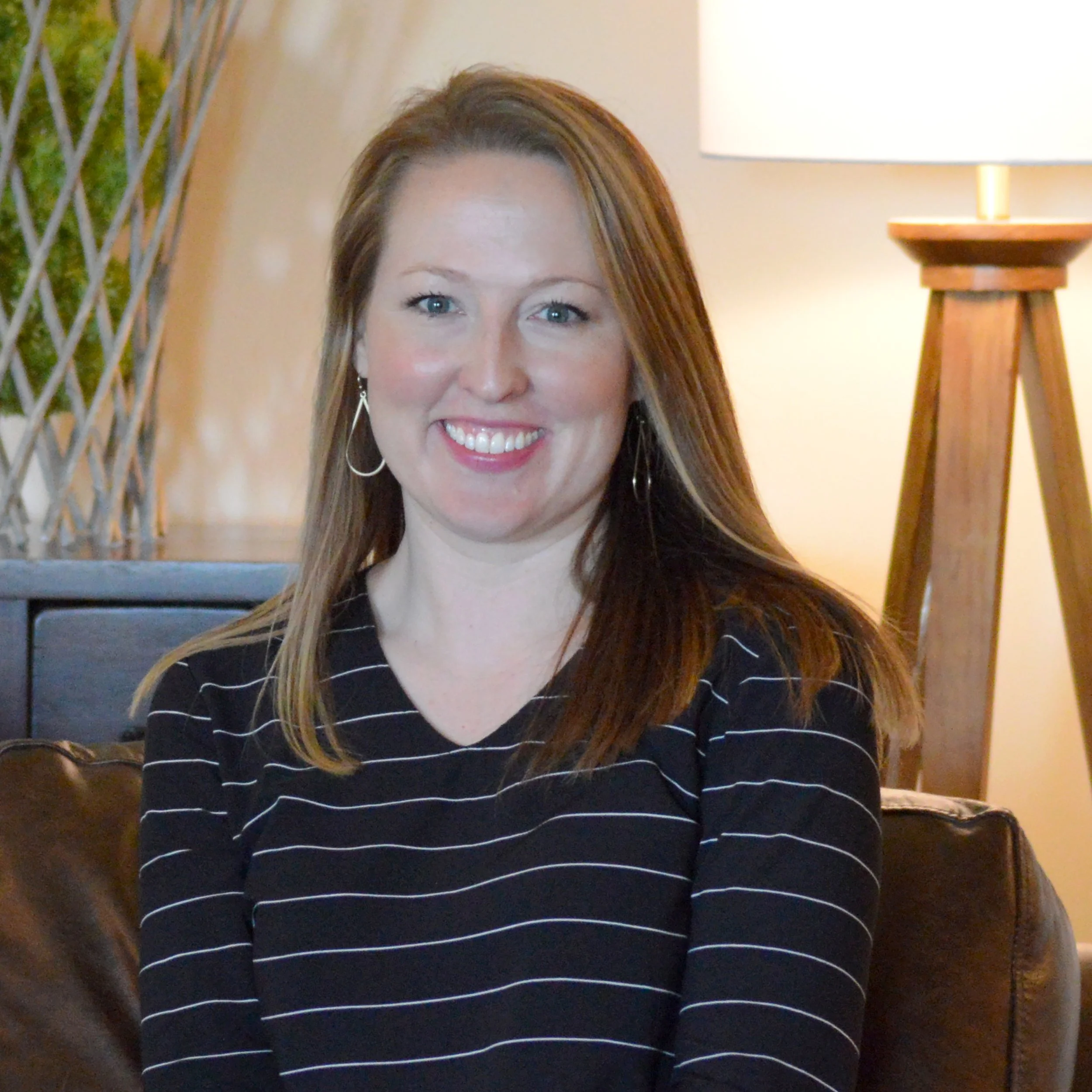 A smiling woman with long straight hair, wearing a black shirt with white horizontal stripes, sitting on a brown leather sofa in a cozy room with a lamp and decorative elements in the background.